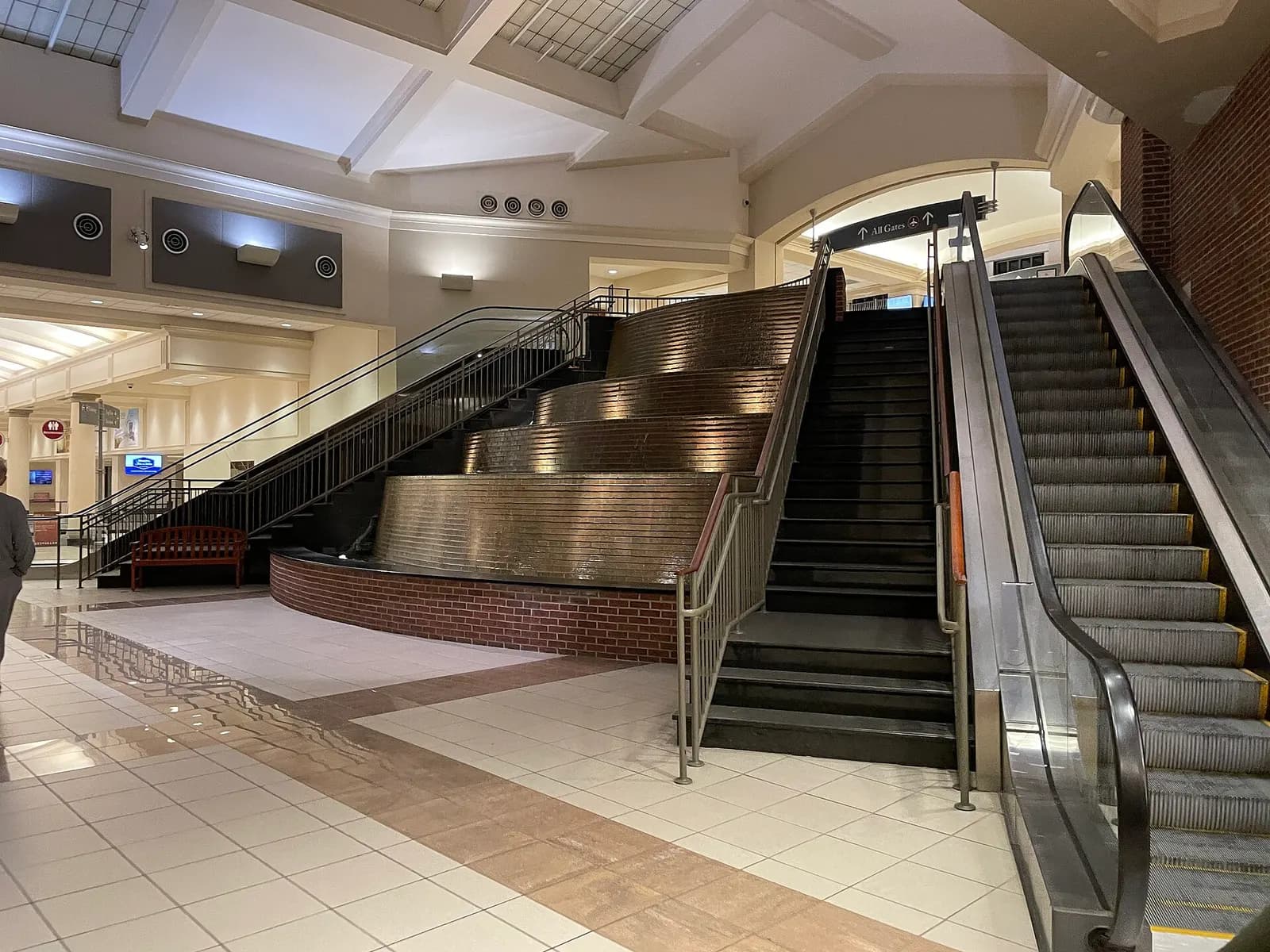 Landside hall with a curved indoor fountain beside stairs and an All Gates escalator at Alexandria International Airport.