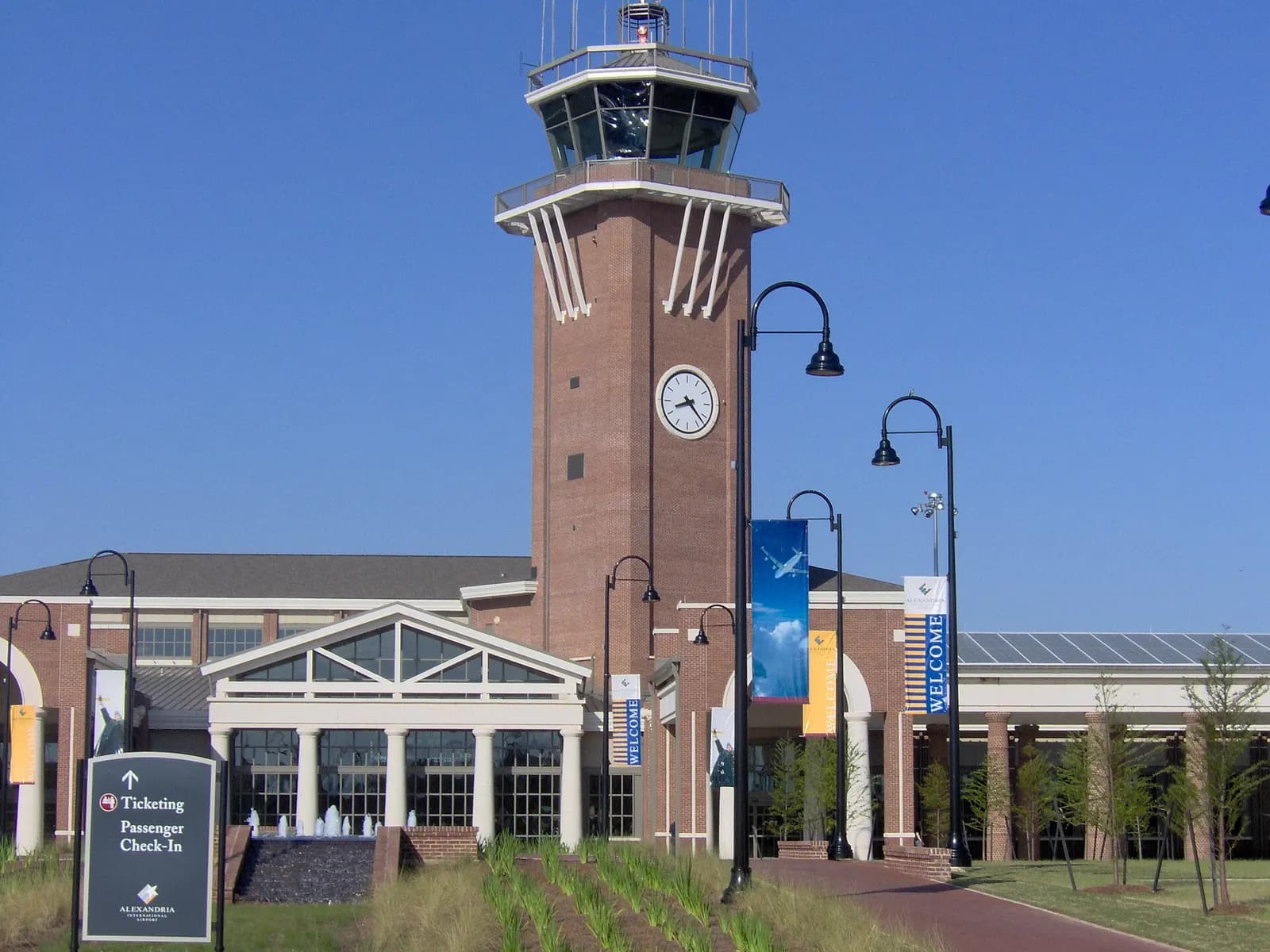 Daytime brick passenger hall and control tower at Alexandria International Airport, with a Ticketing and Passenger Check-In sign on the front lawn.