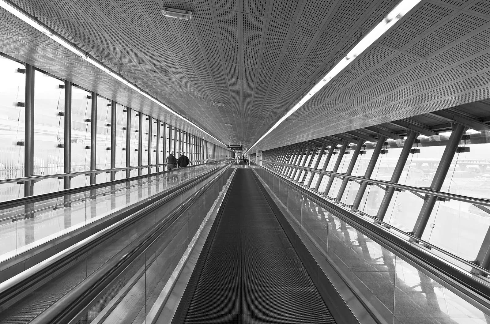 A long glass-walled transfer corridor at Málaga Airport connects terminals, with moving walkways and passengers pulling wheeled luggage.