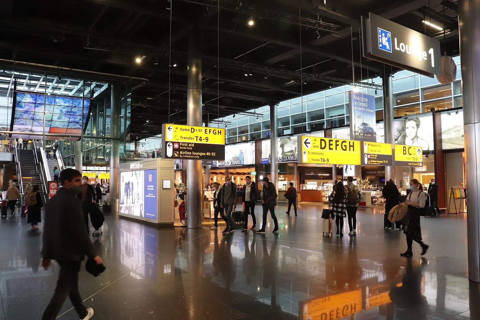 A spacious departures terminal at Schiphol with check-in counters, flight information displays, and passengers moving through the hall.