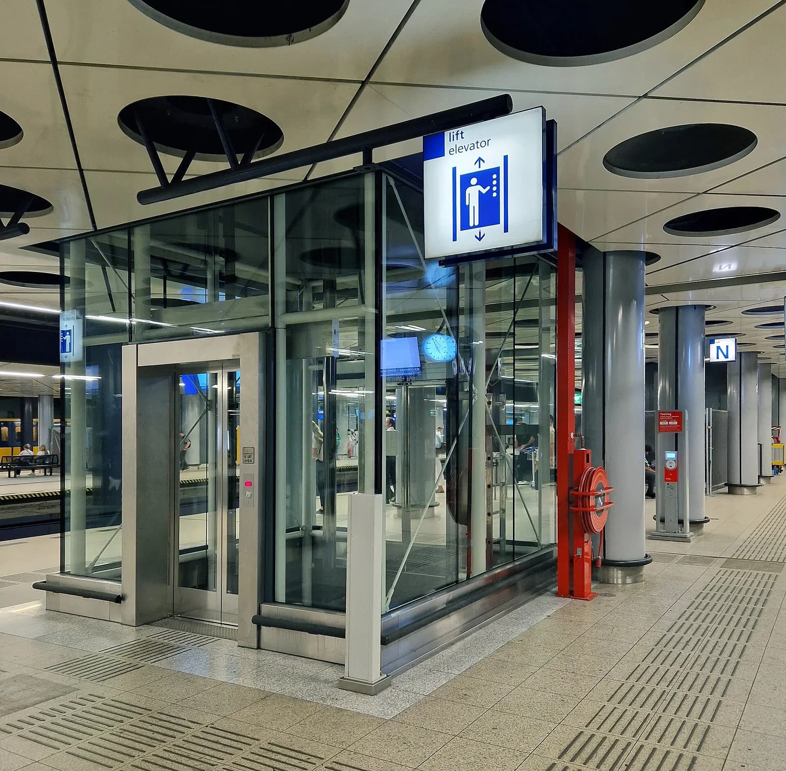 An elevator inside the Schiphol Airport train station, with platform signs and rail connections visible nearby.