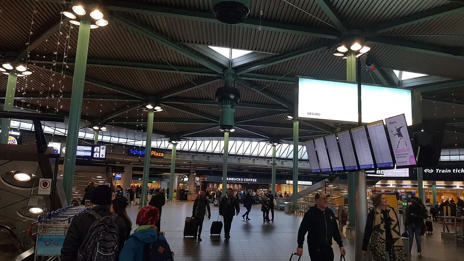 The bright, open Schiphol Plaza hall with shops, cafes, and travelers passing through beneath a high ceiling.