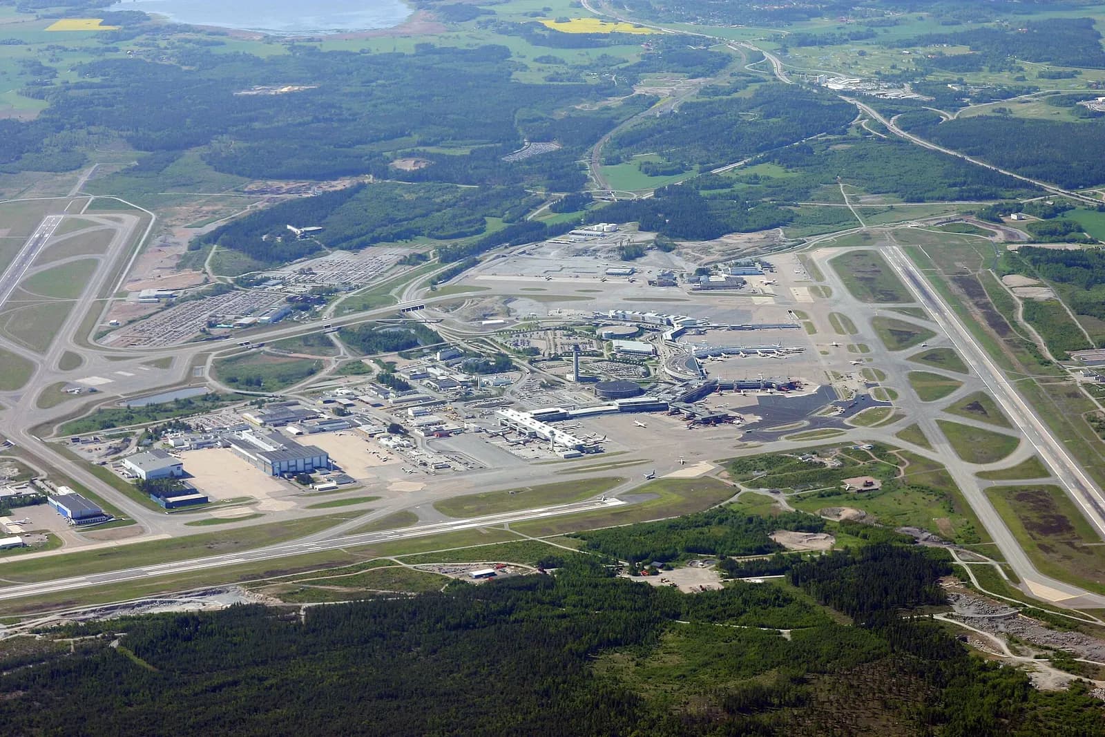 Wide aerial view of Stockholm Arlanda Airport from the north, showing multiple terminals, runways, and surrounding forest.