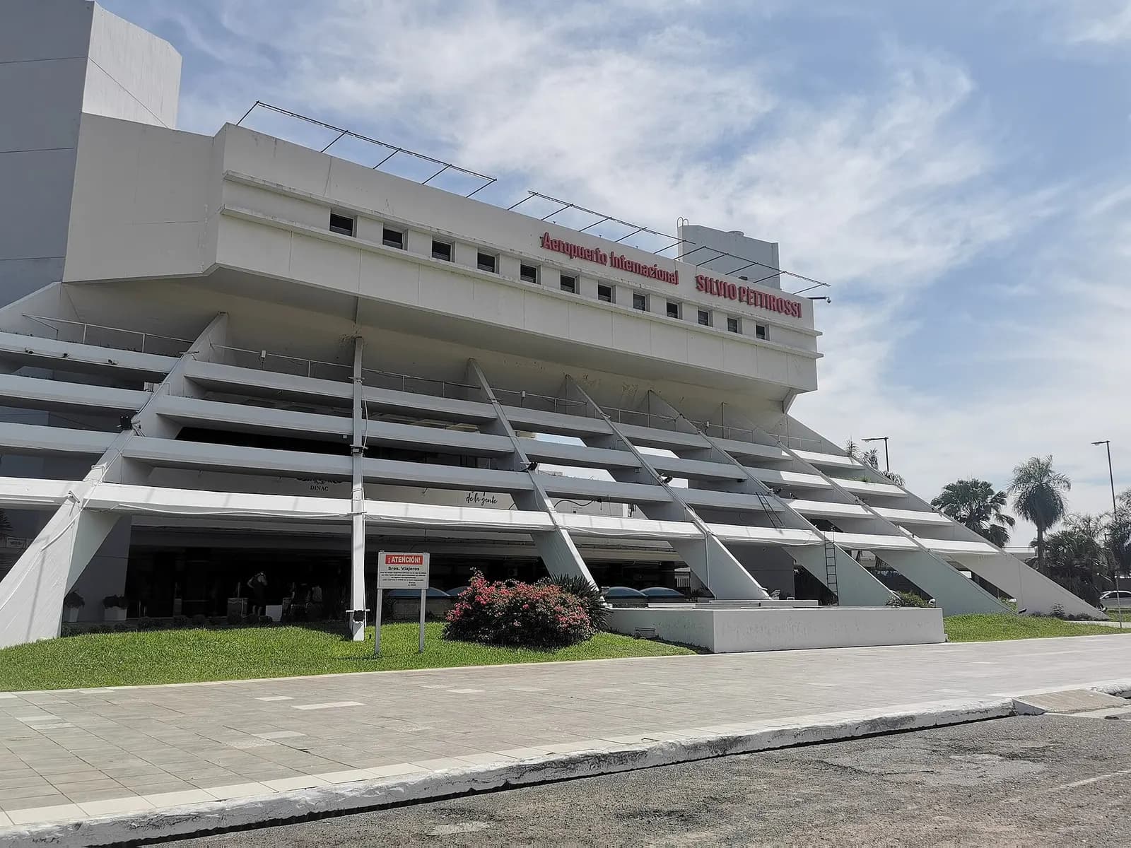 Street-level view of the white multi-level terminal facade with red “Aeropuerto Internacional Silvio Pettirossi” lettering, diagonal ramps, and the curbside entrance.
