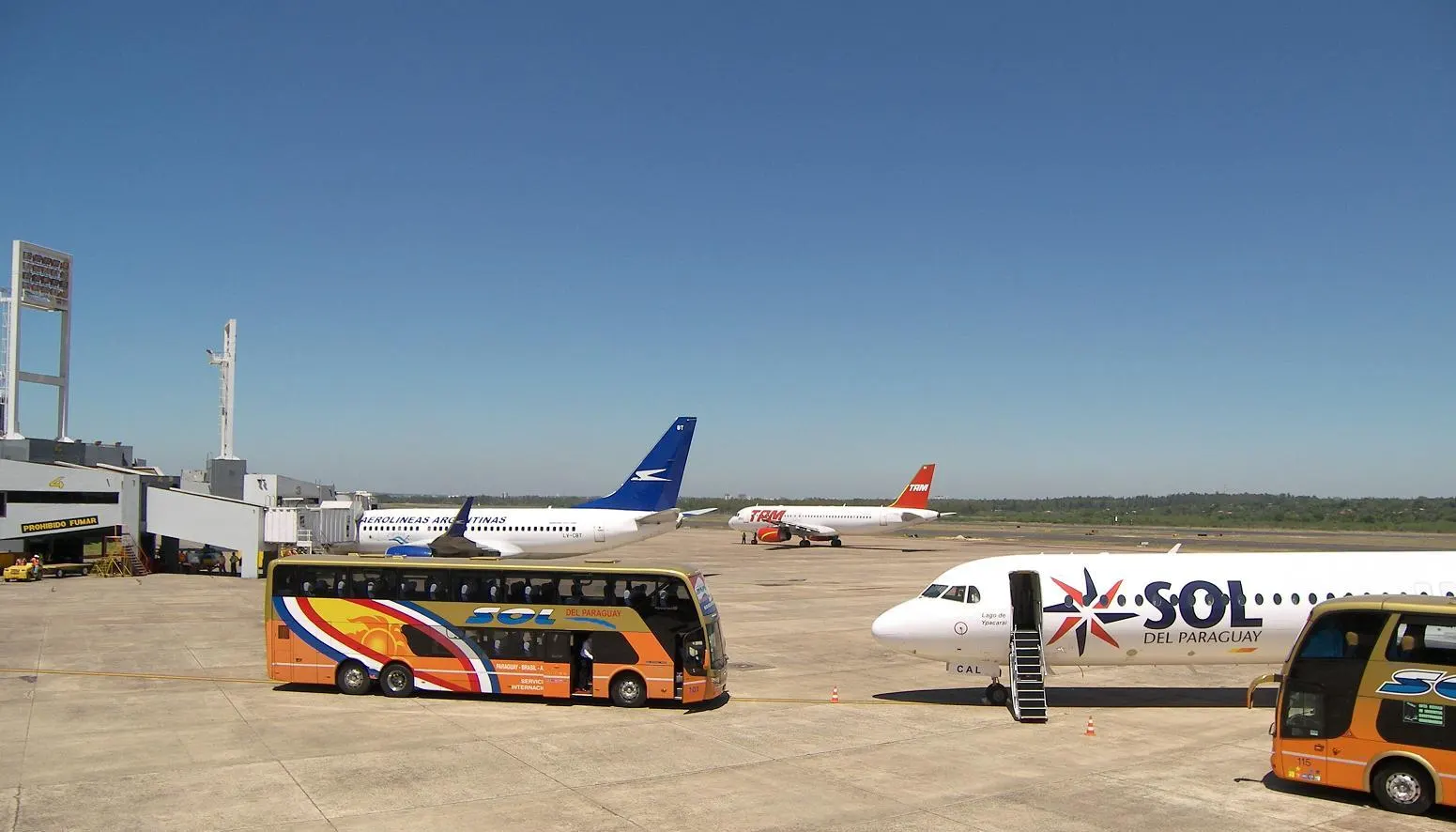 Airport apron under a deep blue sky with a Sol del Paraguay jet and boarding buses in the foreground and other airliners parked near a jet bridge.