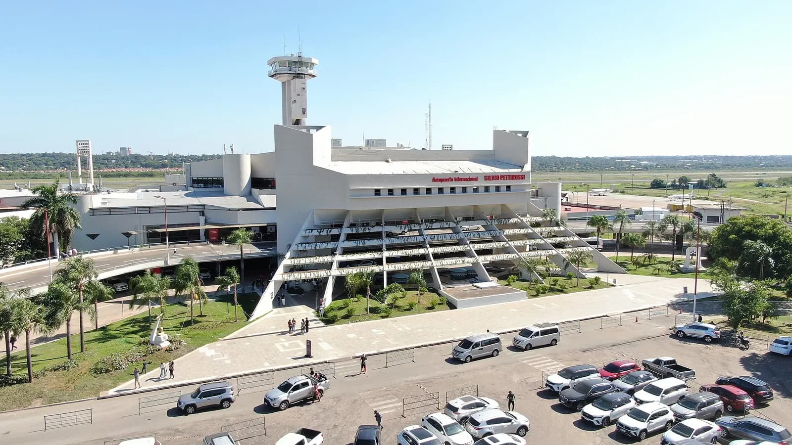 Aerial view of the white stepped main terminal and control tower at Silvio Pettirossi International Airport, with parking lot, palm trees, and roadway in front under a clear sky.