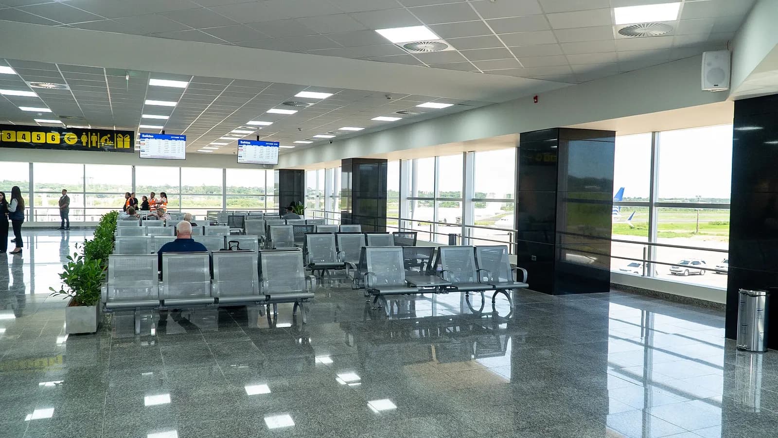 Bright departure lounge with rows of silver metal seats, departure boards, and large windows looking out over the apron and a parked airliner.