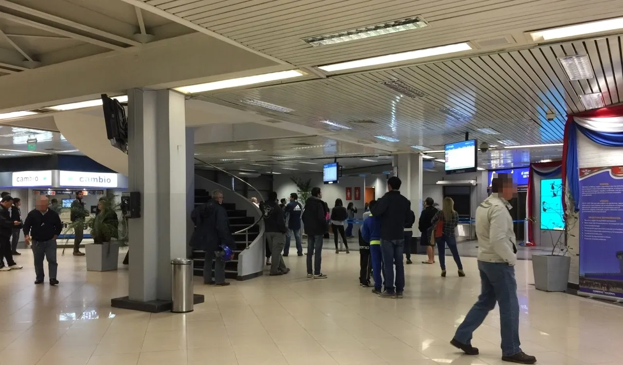 Busy arrivals hall with travelers waiting near a staircase, digital screens, and illuminated “Cambio” currency-exchange counters under fluorescent lighting.
