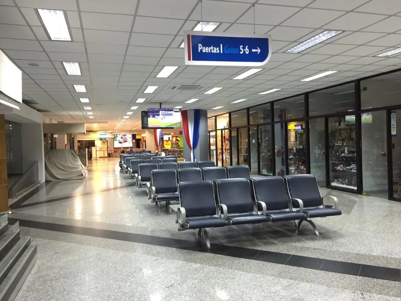 Quiet transit corridor with blue leather gate seats, a hanging “Puertas / Gates 5-6” sign, glass-front retail shops, and Paraguayan flags.