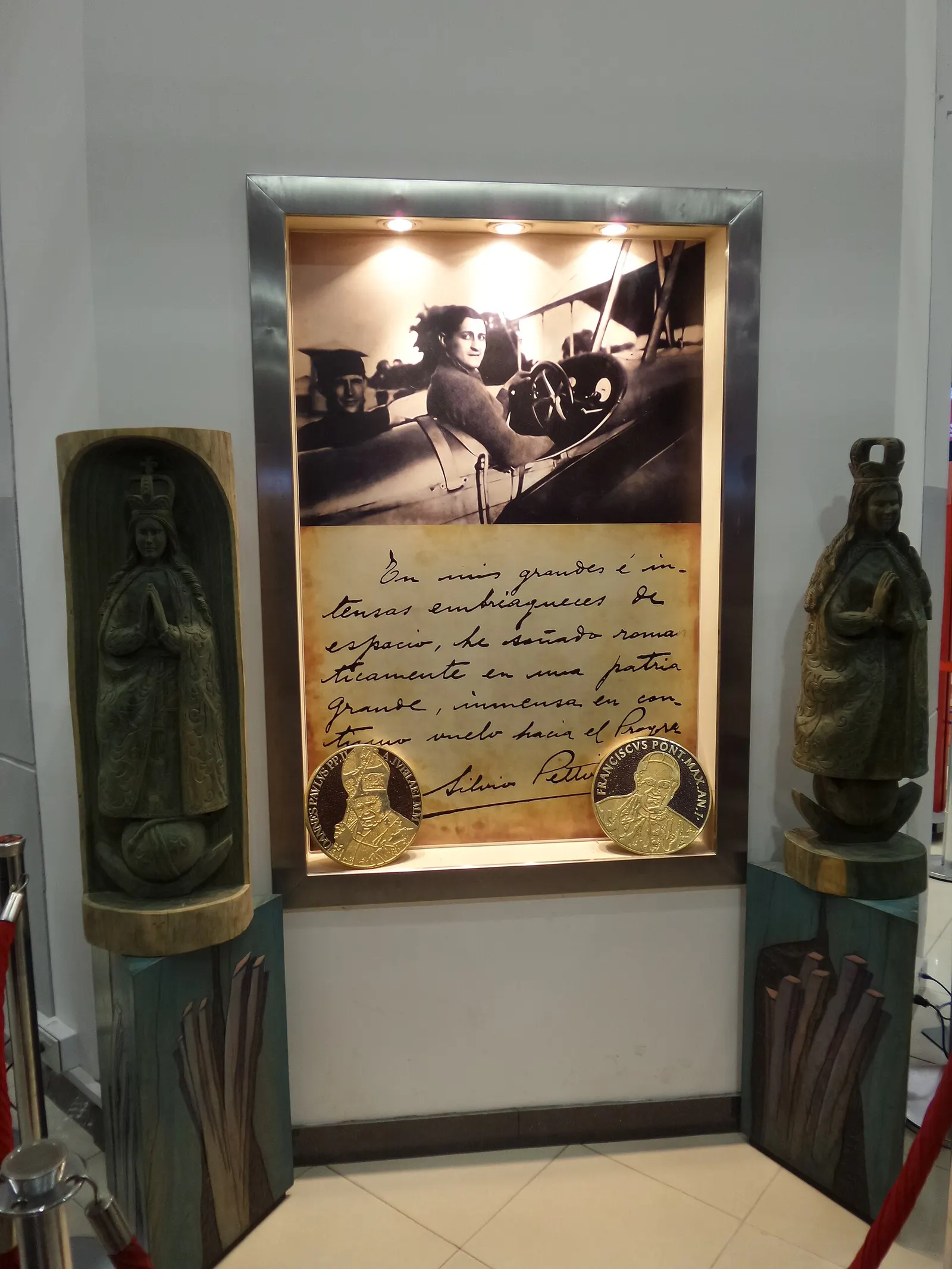 Indoor memorial niche with wooden religious statues flanking a lit display of a historic photo, handwritten note, and medallions dedicated to Silvio Pettirossi.