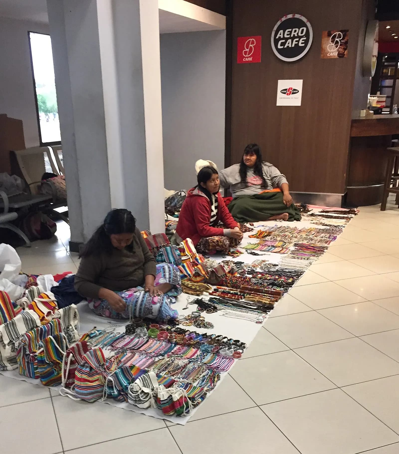 Three women sitting on the terminal floor beside an Aero Café, selling colorful woven bags, textiles, and beaded jewelry laid out on white cloths.