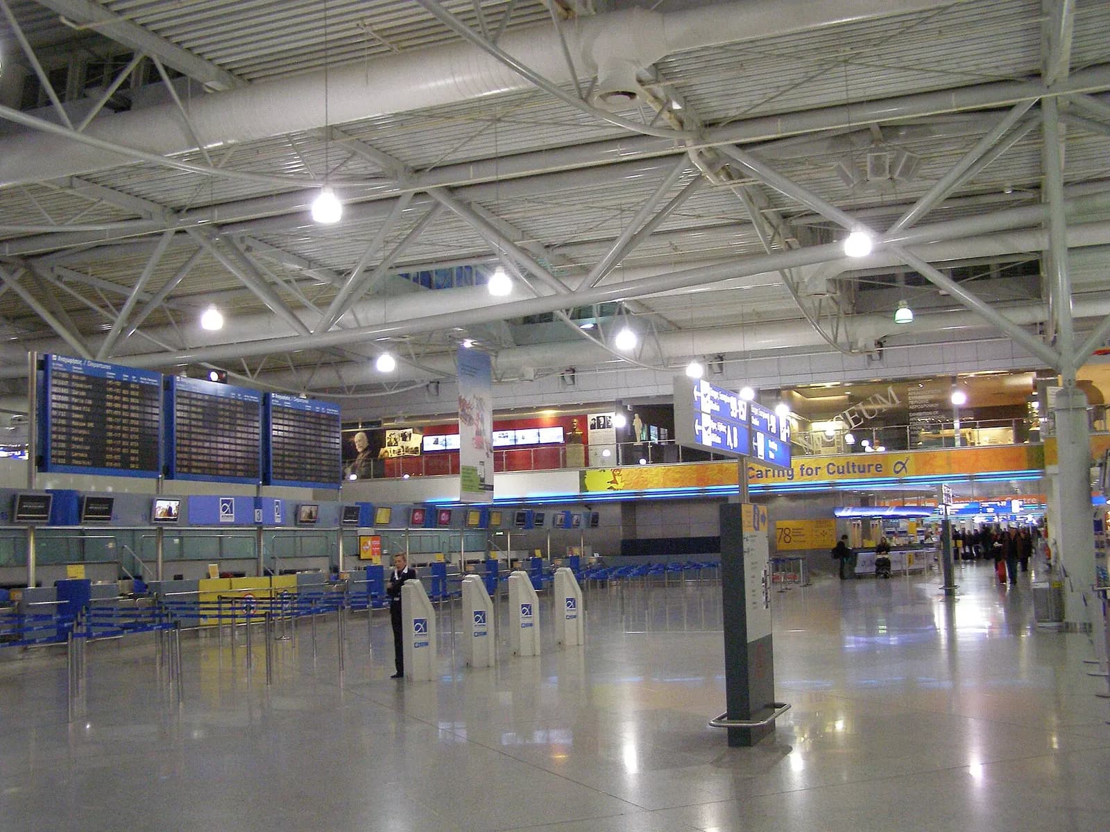 The departure hall at Athens International Airport with check-in areas, flight information screens, and passengers in the open terminal space.