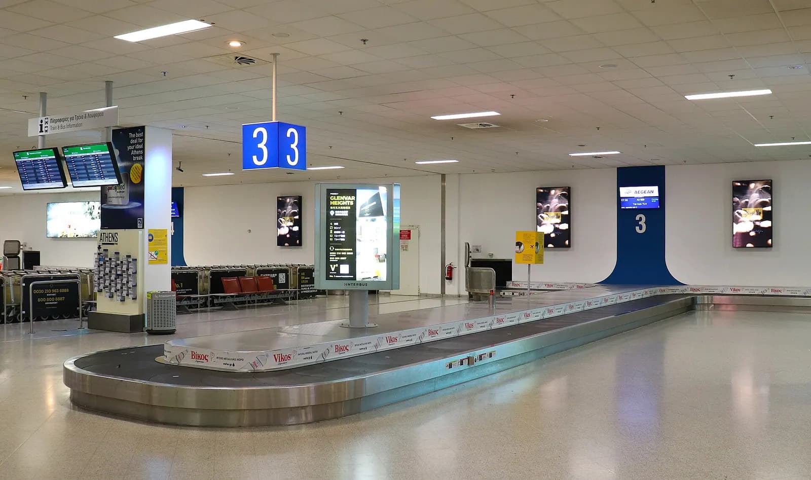 A baggage reclaim carousel in the arrivals hall at Athens International Airport, with overhead signs and an empty conveyor belt.