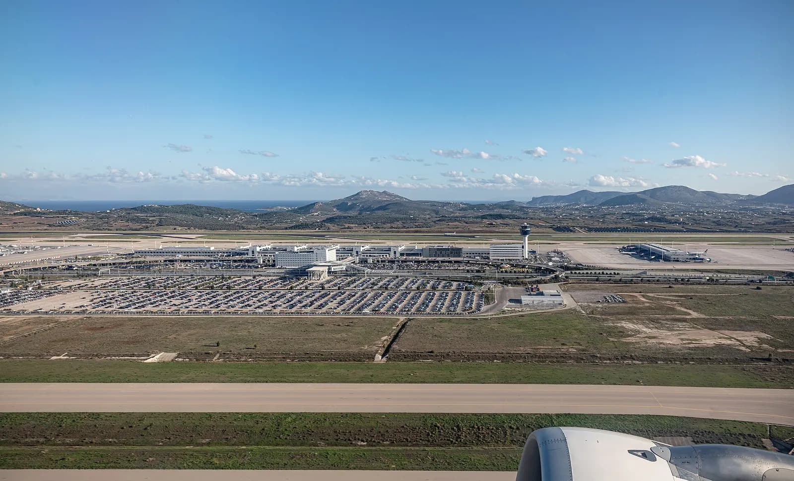 A wide exterior view of the Athens International Airport terminal building with glass walls, entrance roads, and the control tower visible in the background.