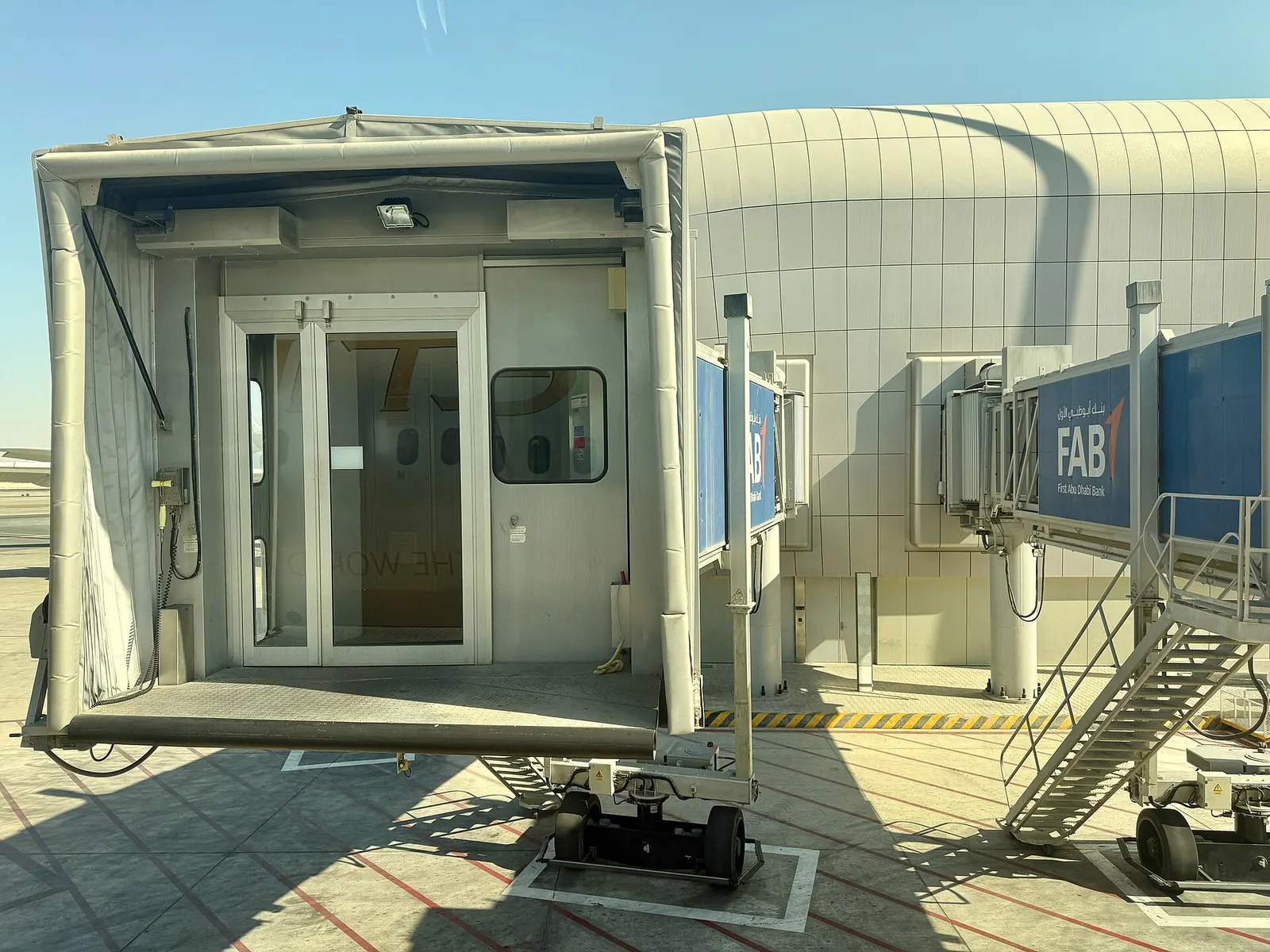 A modern glass jet bridge connected to an aircraft gate at Zayed International Airport, seen from the terminal side on the apron.