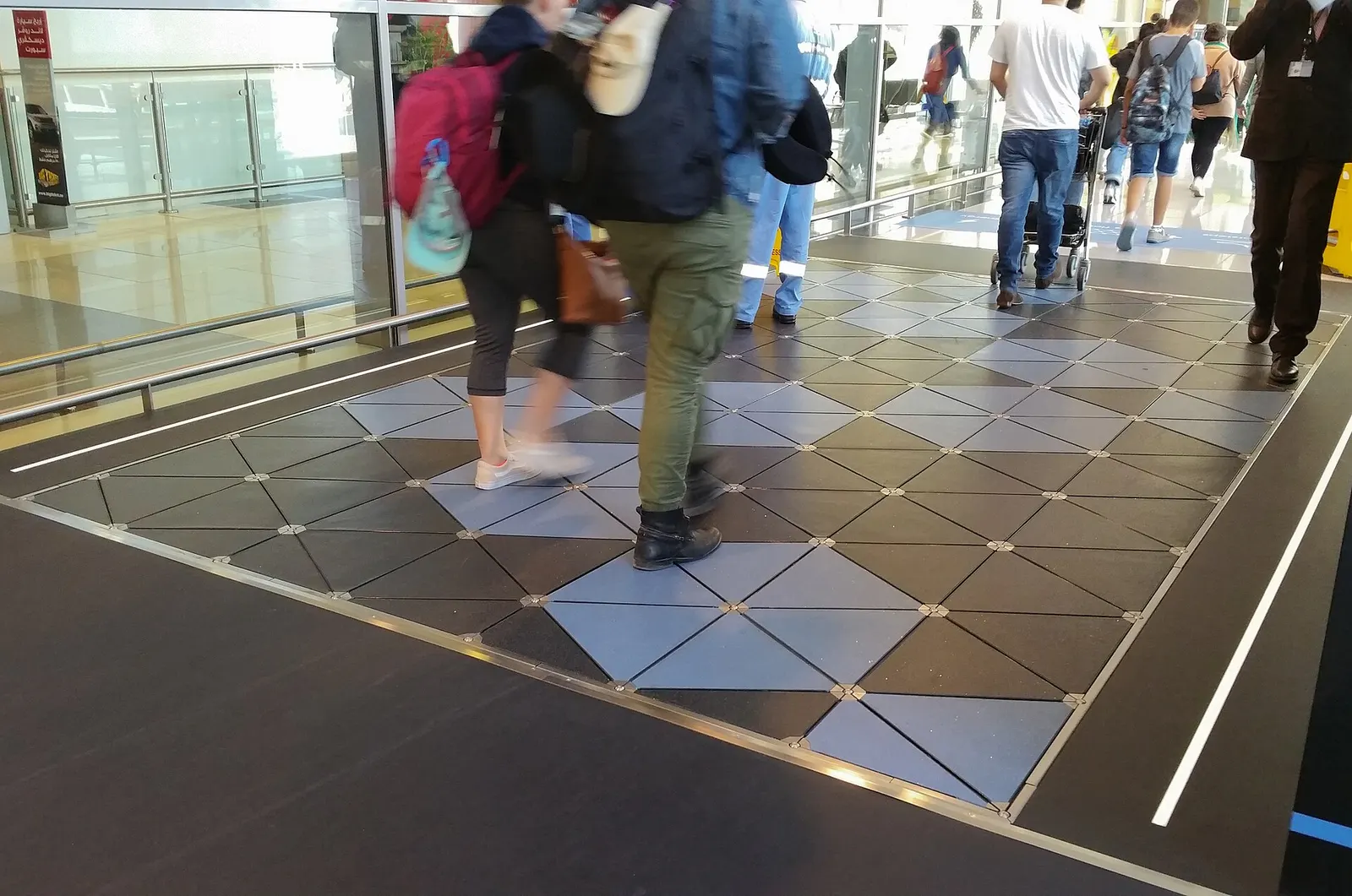 Passengers and airport staff walking across illuminated Pavegen floor tiles in a terminal corridor that converts footsteps into electricity.
