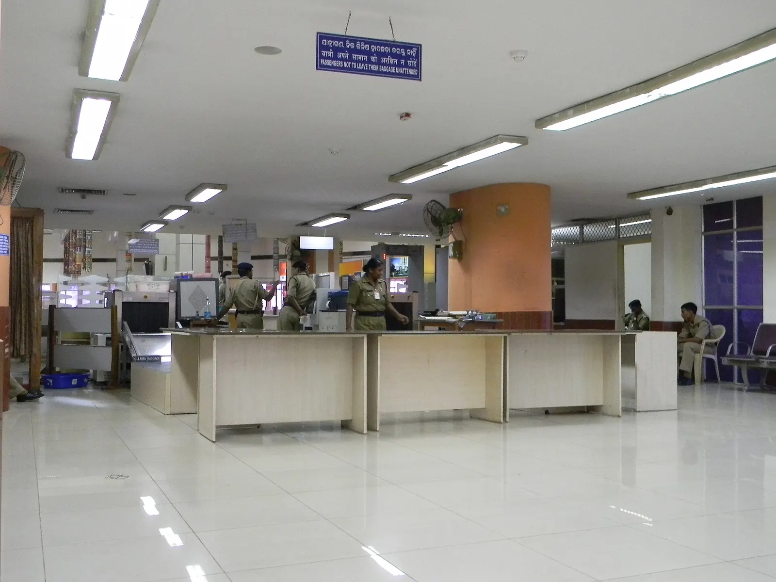 CISF baggage screening tables and X-ray lane inside Biju Patnaik Airport, with Odia/Hindi/English unattended-baggage signage