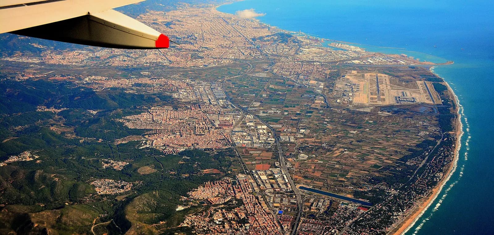 Aerial view of Barcelona-El Prat Airport south of the city, with Barcelona's port, Montjuïc hill, and urban coastline visible in the background.