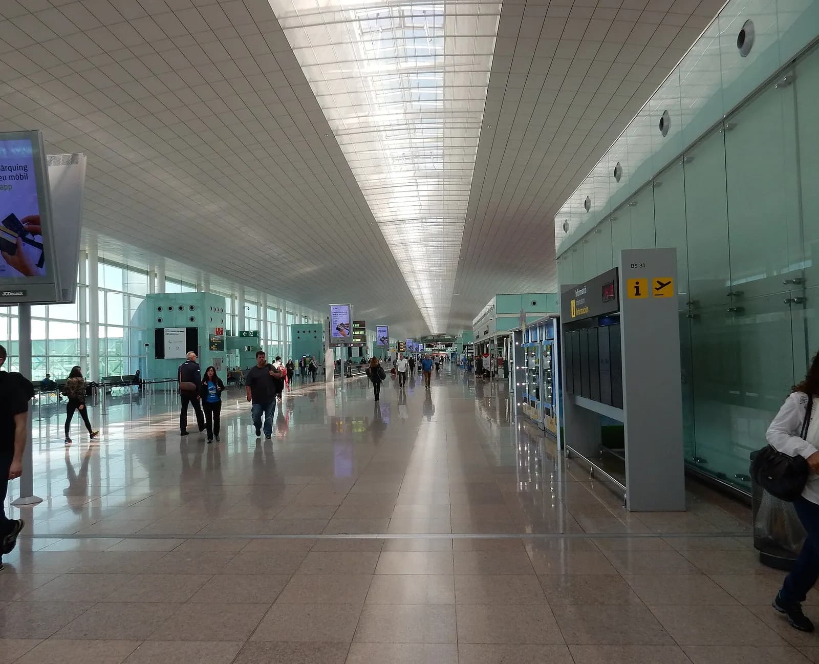 Bright, spacious Terminal 1 concourse at Barcelona-El Prat with high ceilings, large windows, shops, and seating areas.