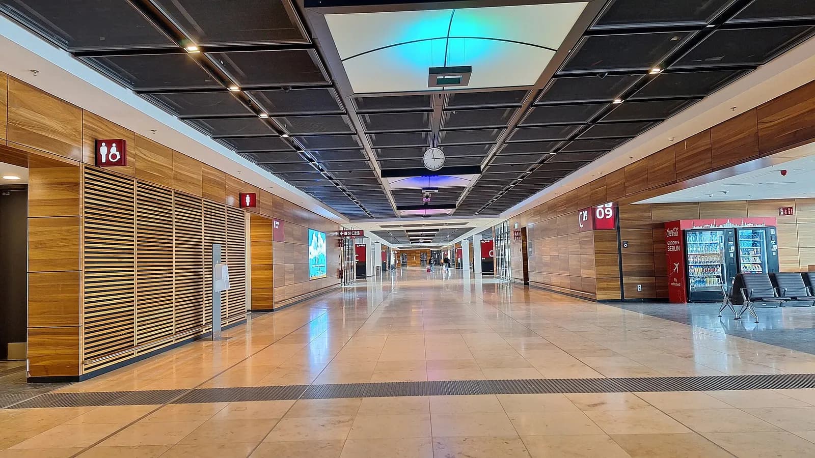 The boarding area at Berlin Brandenburg Airport has rows of gate seating, overhead flight displays, and large windows facing the apron.