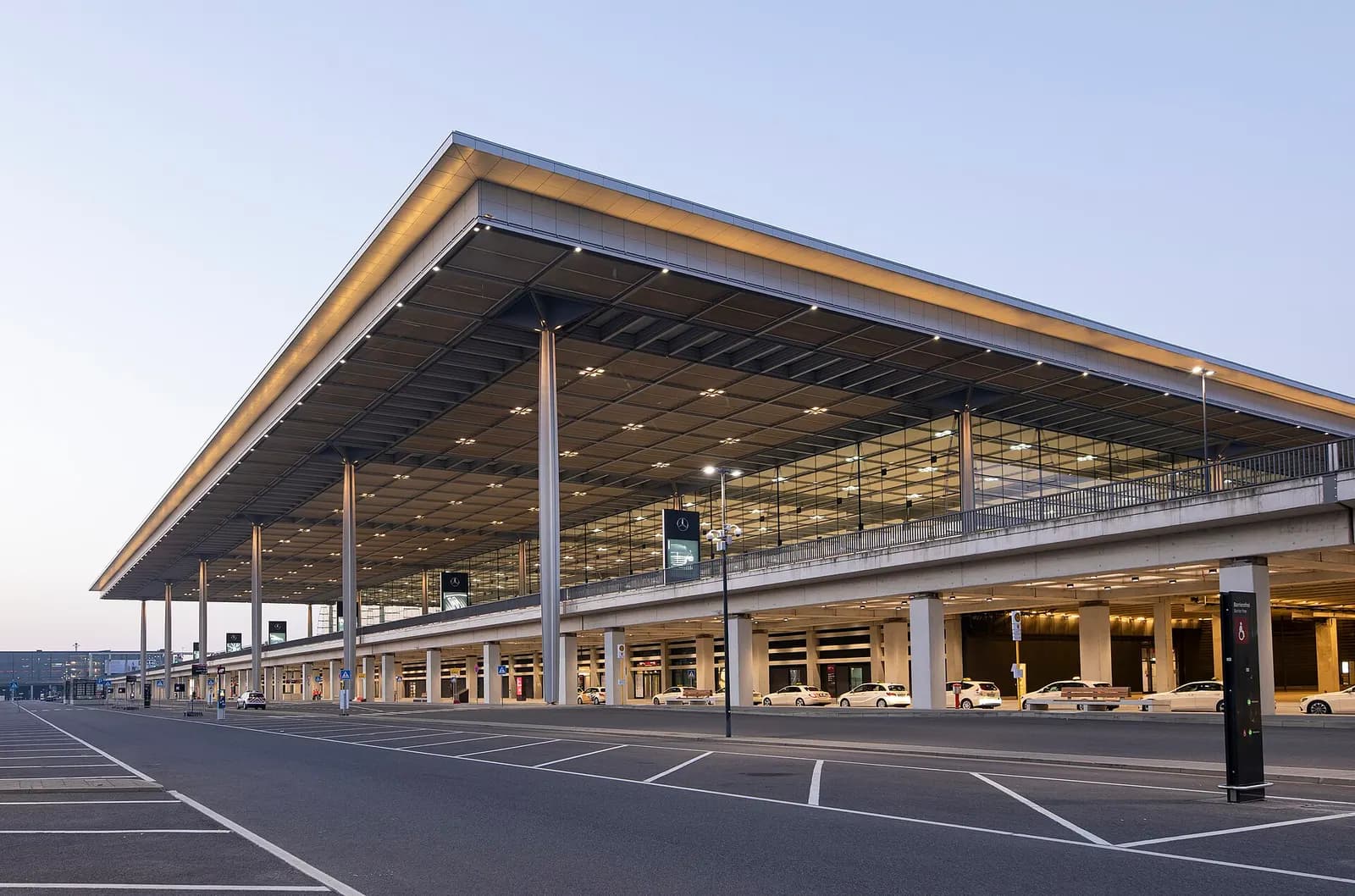 The glass-fronted facade of Berlin Brandenburg Airport Terminal 1 stretches across the frame under daylight, with entrance areas and landscaping in front.