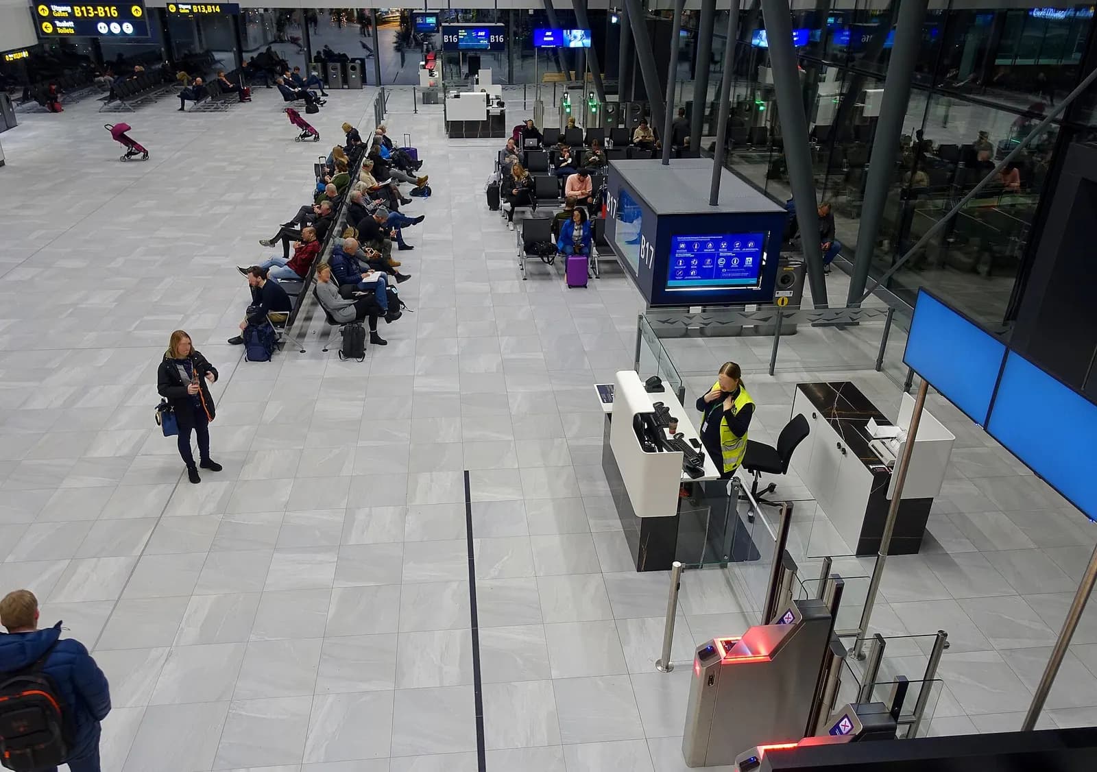 Seated pasengers waiting before boarding their flight, by the gates B16 and B17 in the departure hall in the terminal of Bergen Airport, Flesland , an international airport located outside Bergen, Norway, on November 21st, 2019. Airport employee in reflective vest behind the counter. Boarding card r