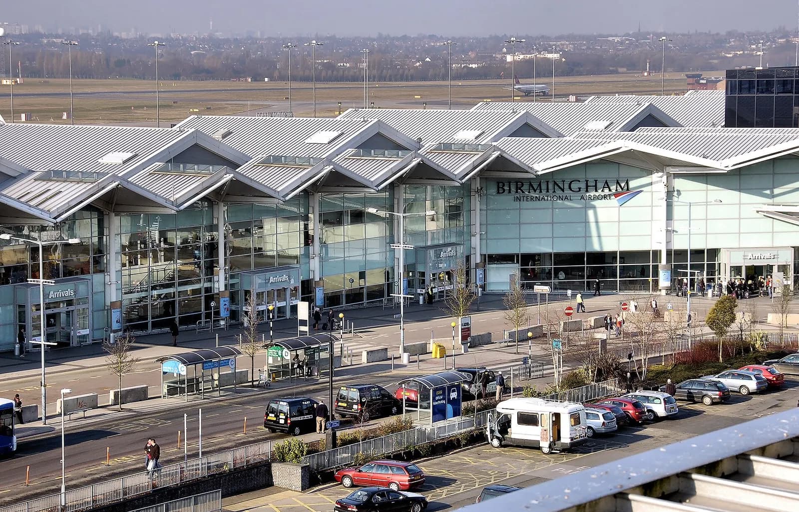 Terminal 2 at Birmingham Airport viewed from a multi-storey car park, with the runway and an aircraft visible beyond the building.