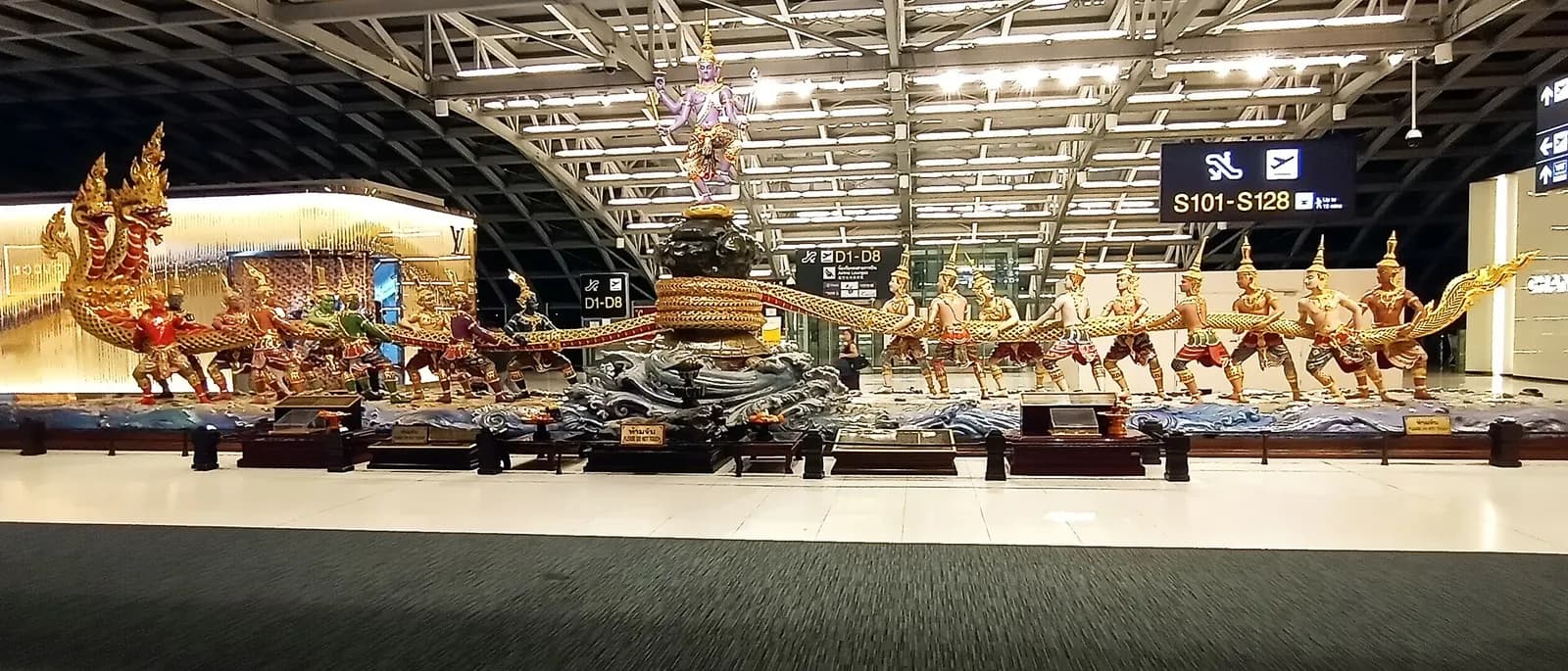 A large bronze sculpture of demons and a serpent depicting the Hindu Churning of the Ocean of Milk, displayed inside the terminal.