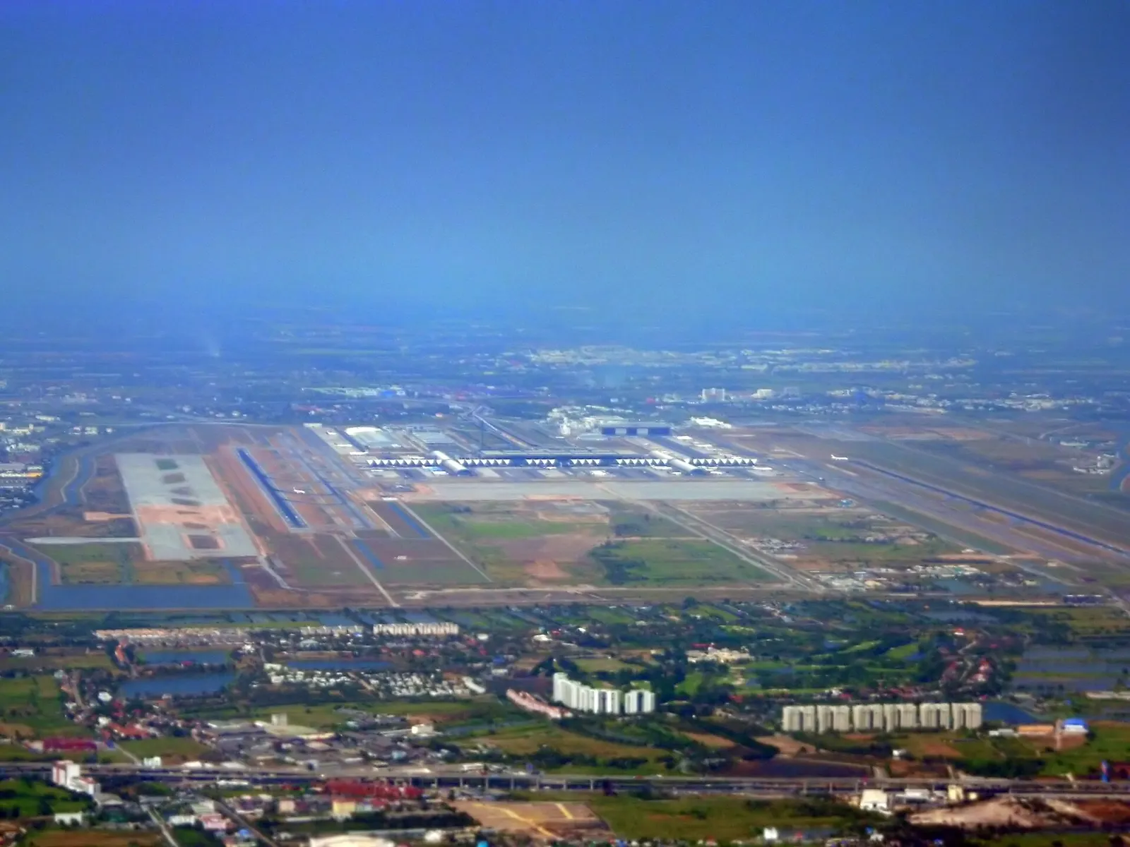 An aerial view of Suvarnabhumi Airport showing the X-shaped terminal, parallel runways, taxiways, and surrounding countryside east of Bangkok.