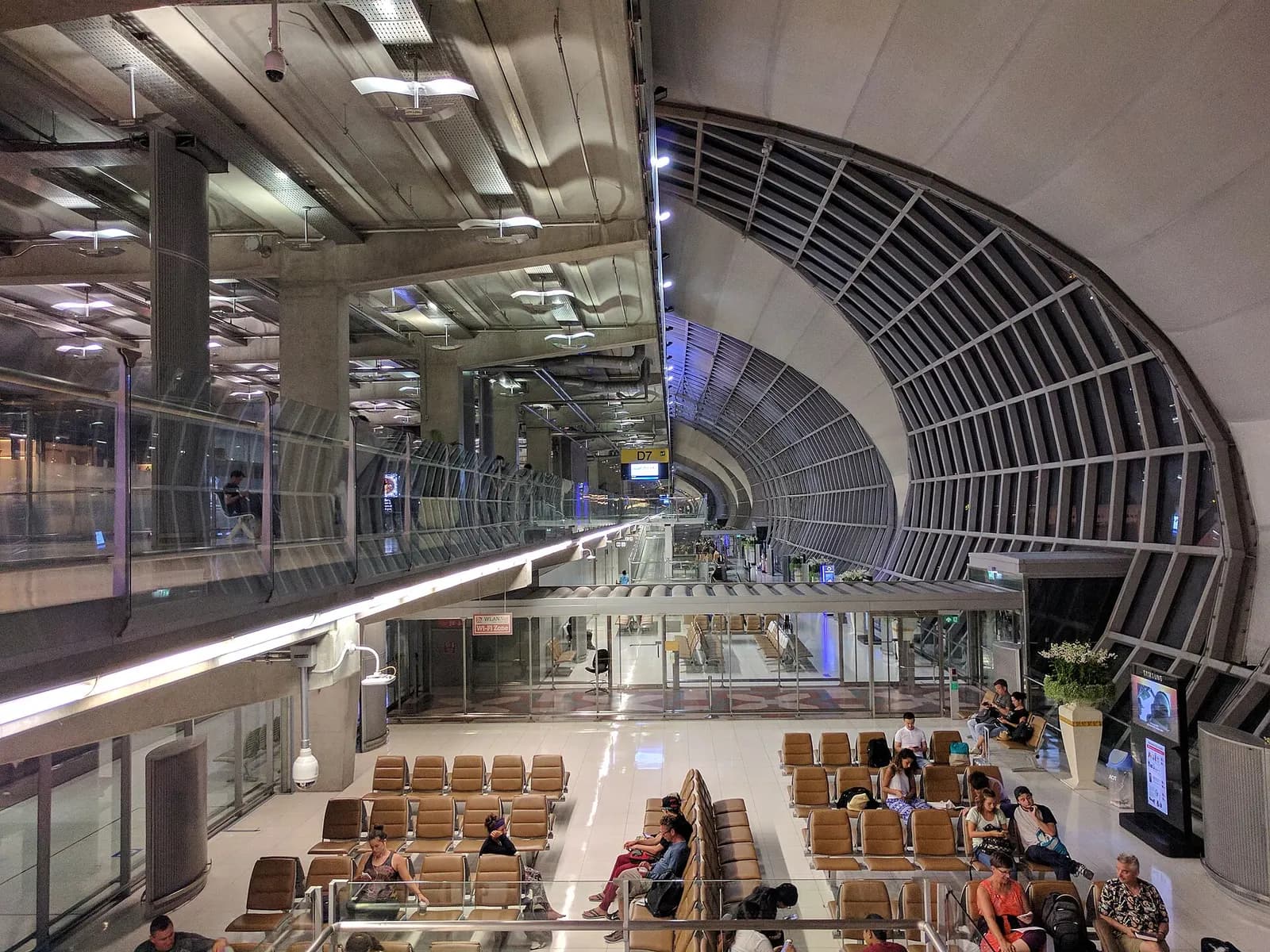 A bright gate waiting area in Terminal D with seating rows, flight-information displays, and passengers near boarding gates seen from above.
