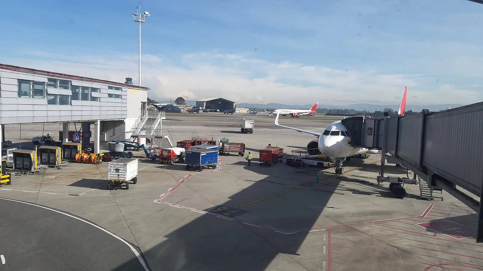 A jet bridge connects a parked airliner to the terminal building on the tarmac, with snow-capped Andes mountains visible in the distance.