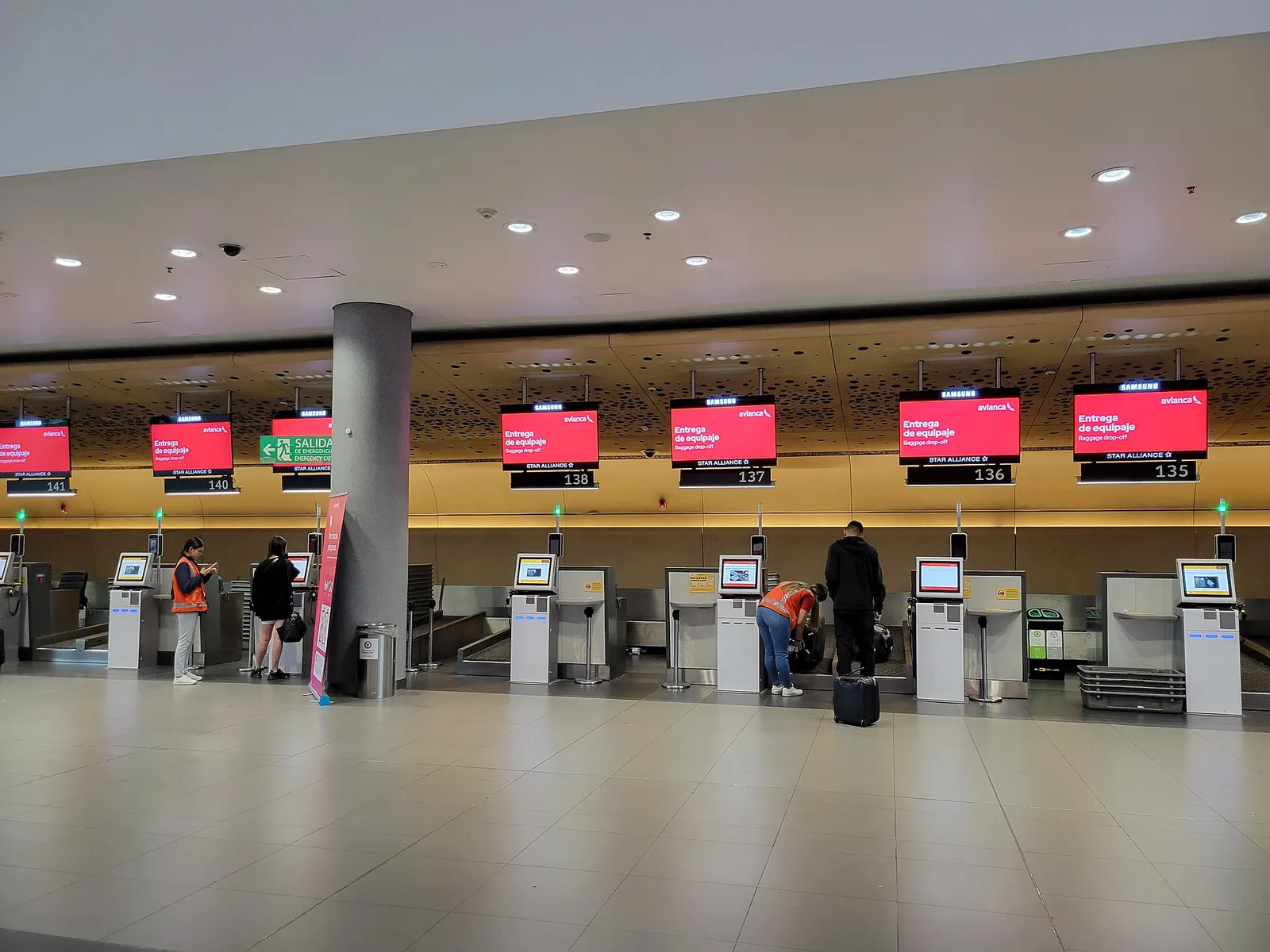 Travelers use a row of self-service check-in kiosks beneath red digital flight information screens in a wood-paneled check-in zone.