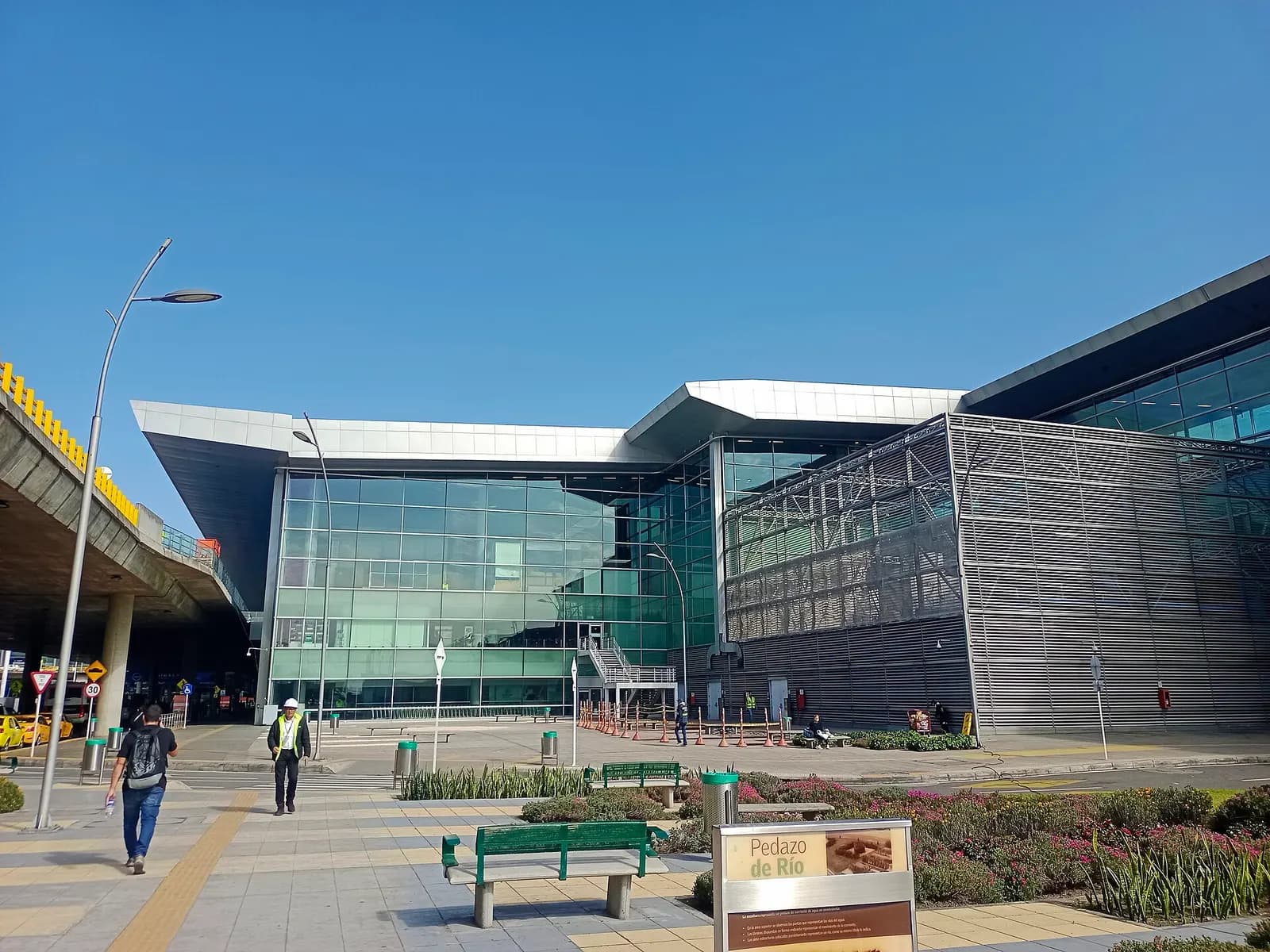 Terminal 1's glass-and-white exterior rises above a landscaped public plaza with green benches under a clear blue sky.