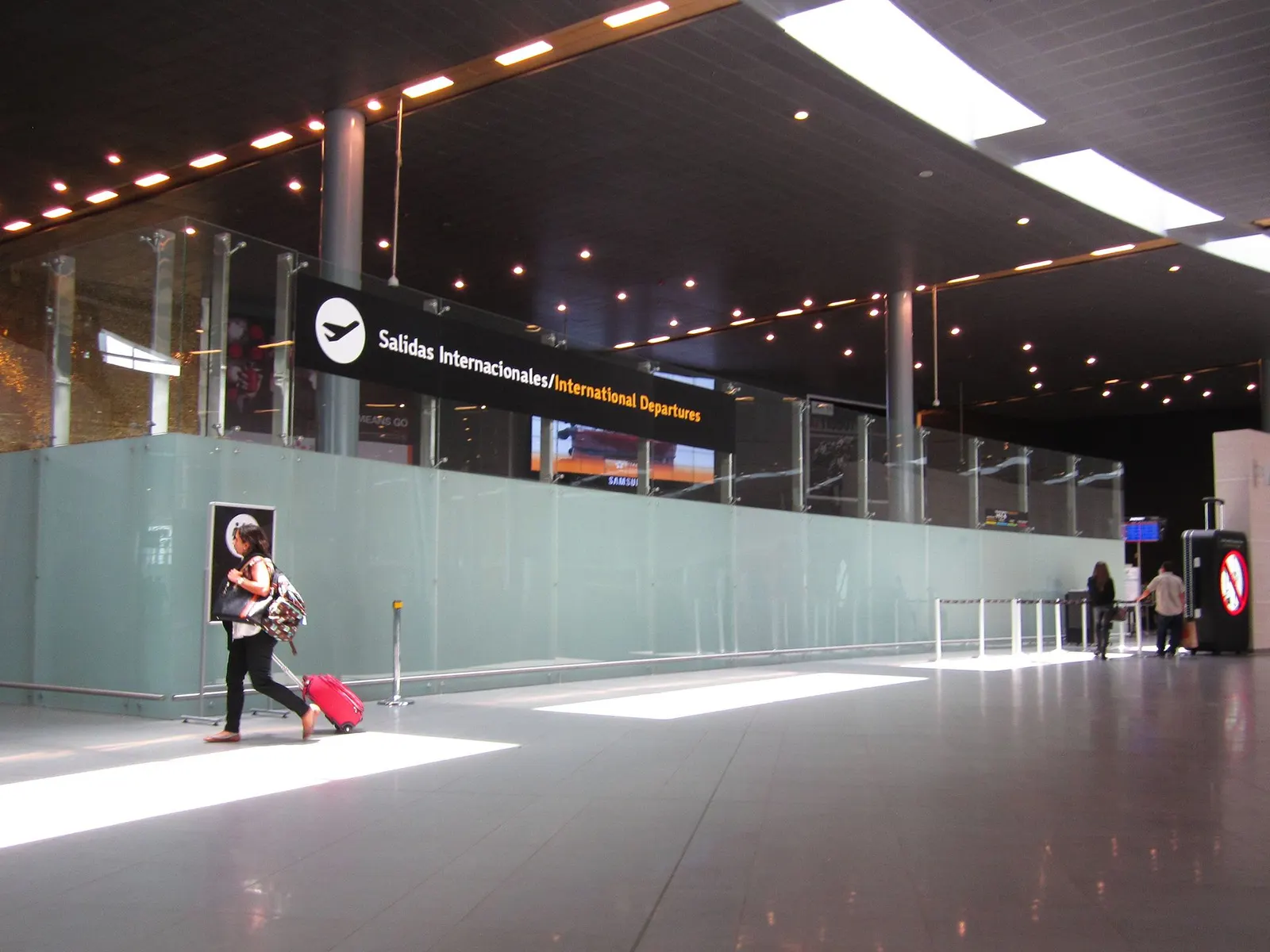 A traveler pulls a suitcase through the international departures hall beneath a bilingual Salidas Internacionales / International Departures overhead sign.
