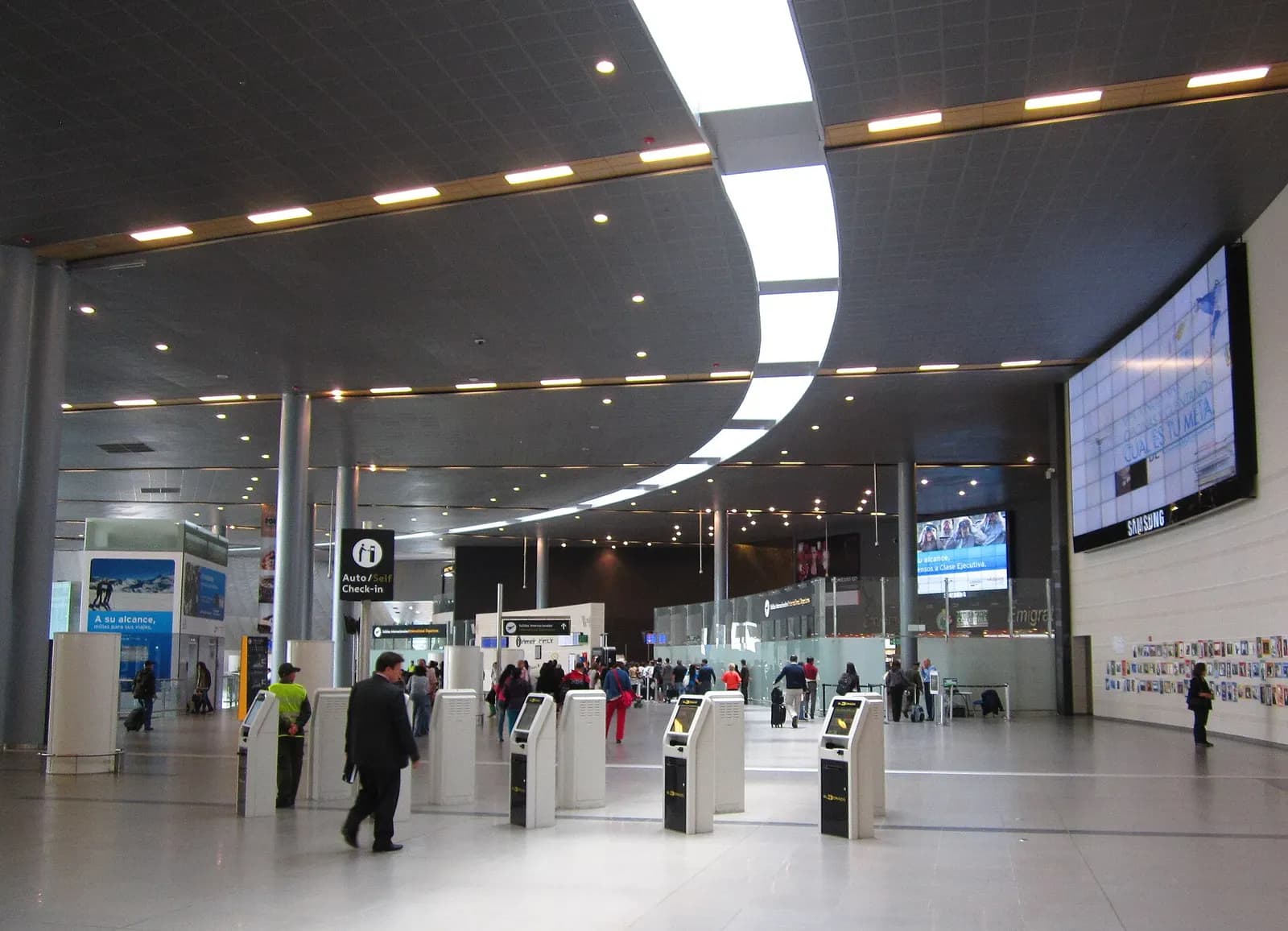 Passengers walk through a wide, modern check-in hall with curved ceiling lighting, white pillars, and self-service kiosks.