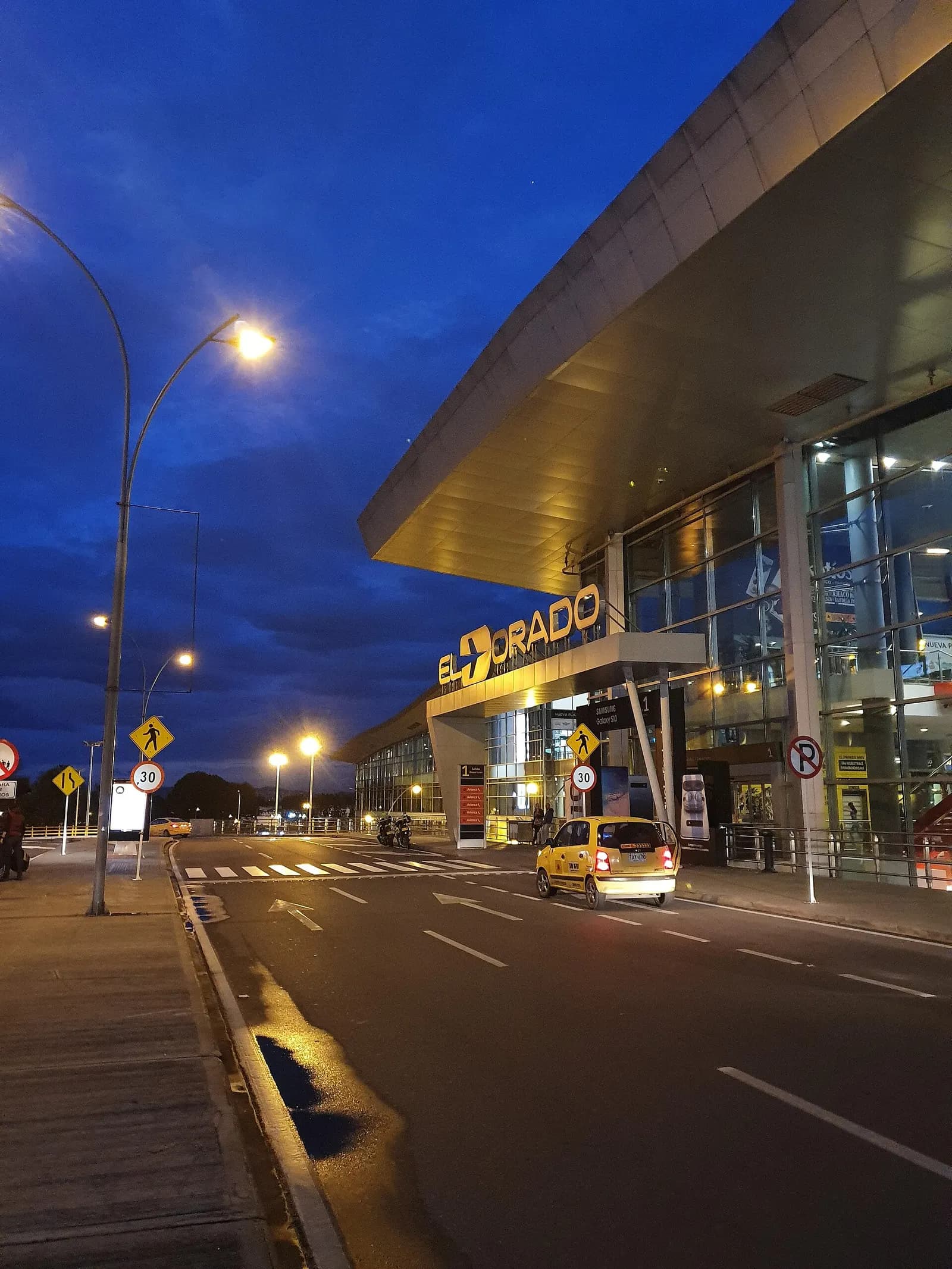 The illuminated EL DORADO sign and glass-fronted main terminal facade at night, seen from the airport access road with a taxi crossing the foreground.