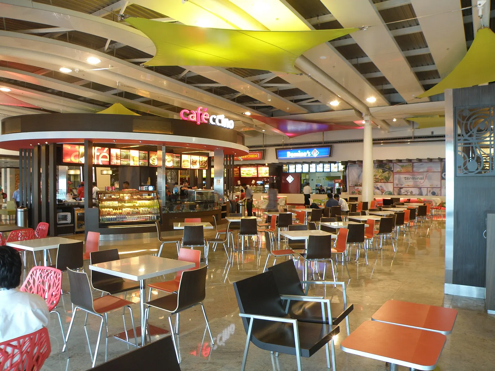 Rows of quick-service restaurants and café seating fill a food court corridor connecting Terminals 1A and 1C.
