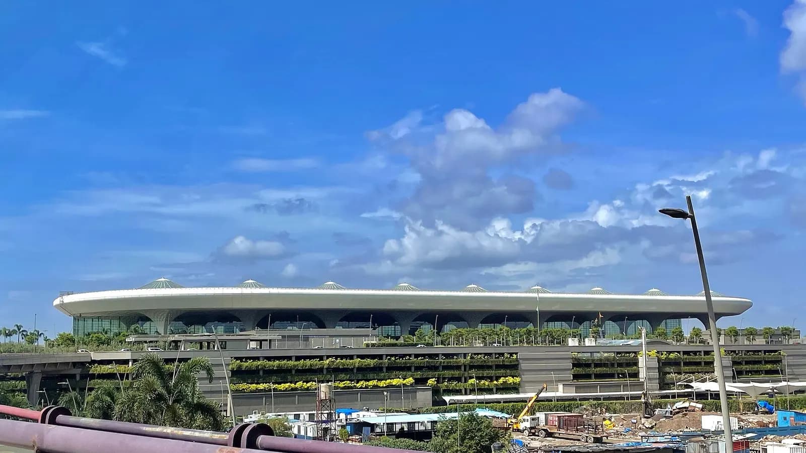 The curved, multi-story Terminal 2 building of Mumbai's airport rises beside the Sahar Elevated Road, with glass facades and landscaped approach roads in the foreground.