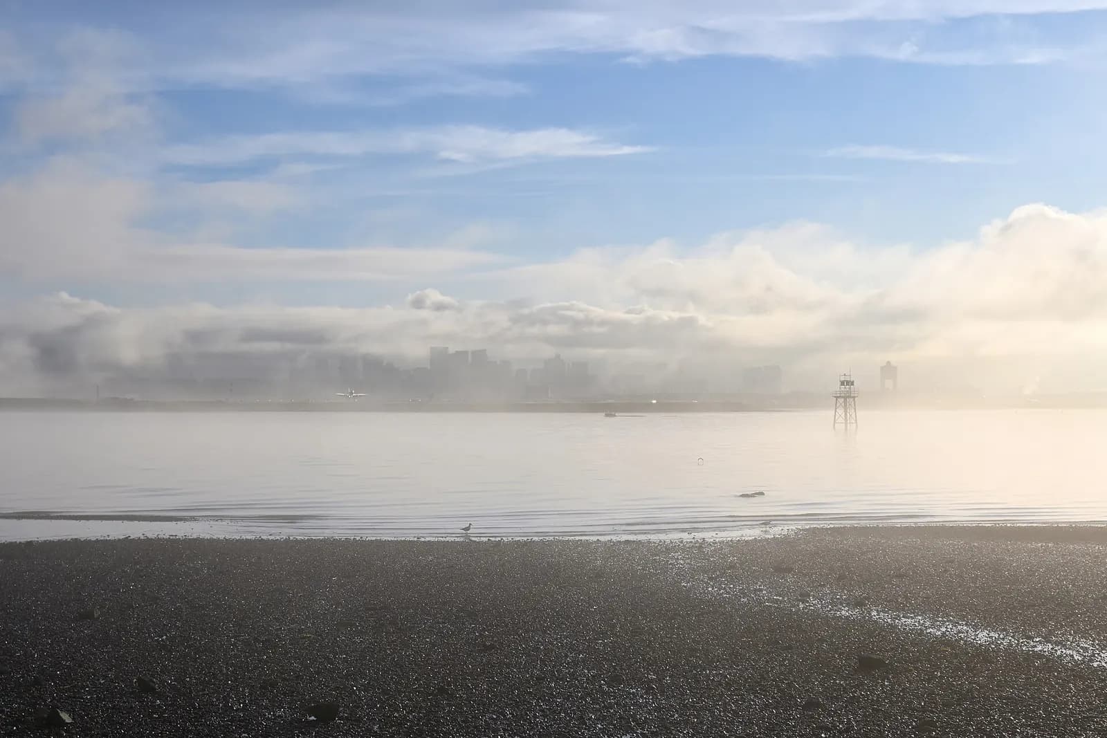 The Boston skyline rises behind Boston Logan International Airport on a foggy day, viewed from Winthrop across the harbor.