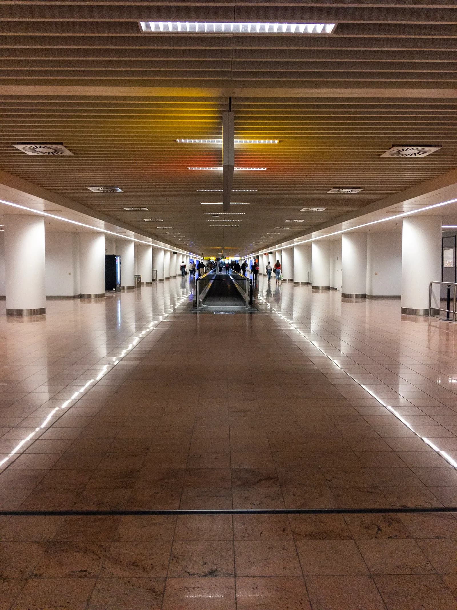 A long indoor passenger corridor at Brussels Airport with ceiling lights, shopfronts, and travelers walking toward the gates.