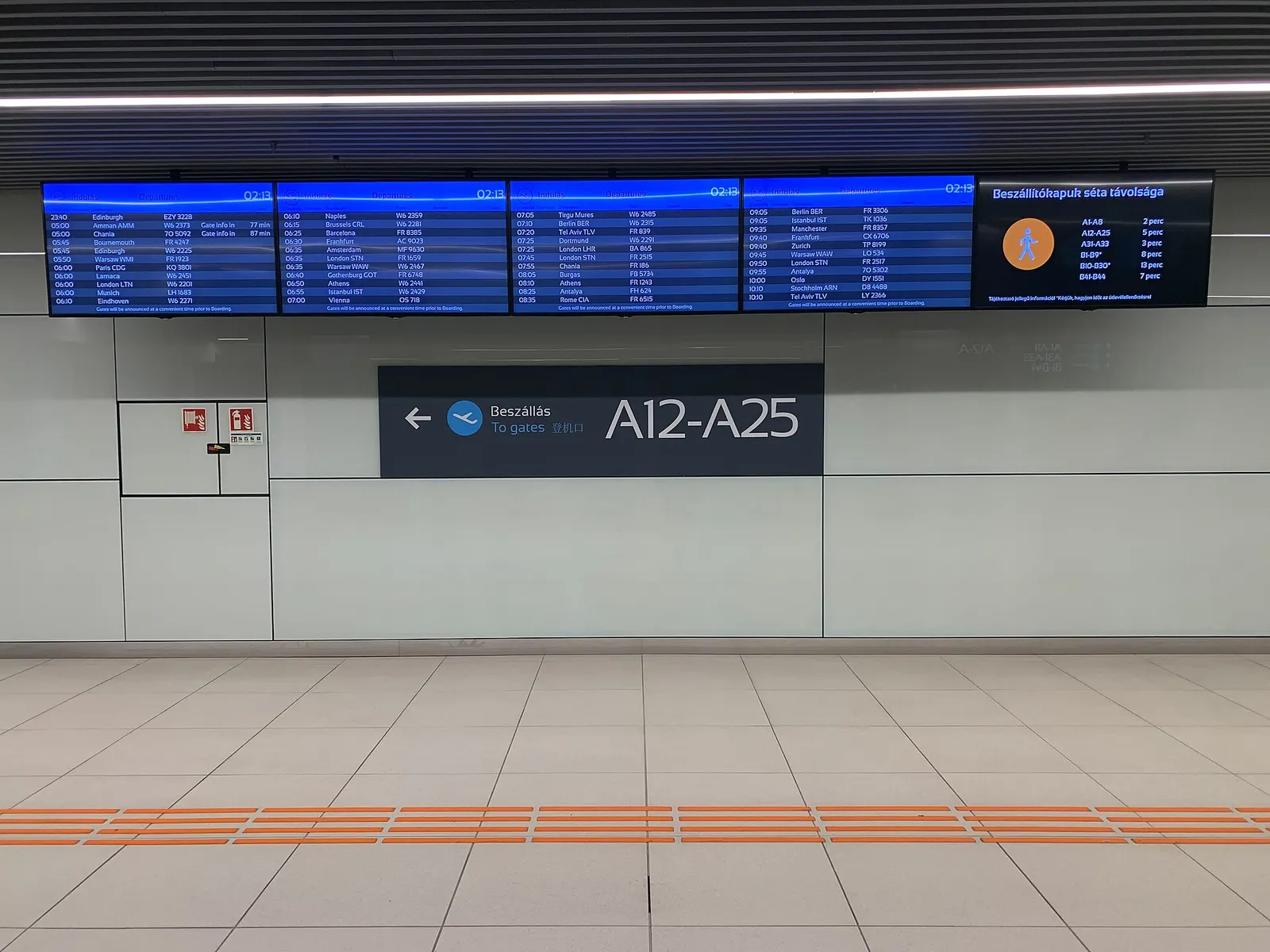 Overhead gate-direction signs and a digital flight-information display in Terminal 2 showing gate numbers and walking distances.