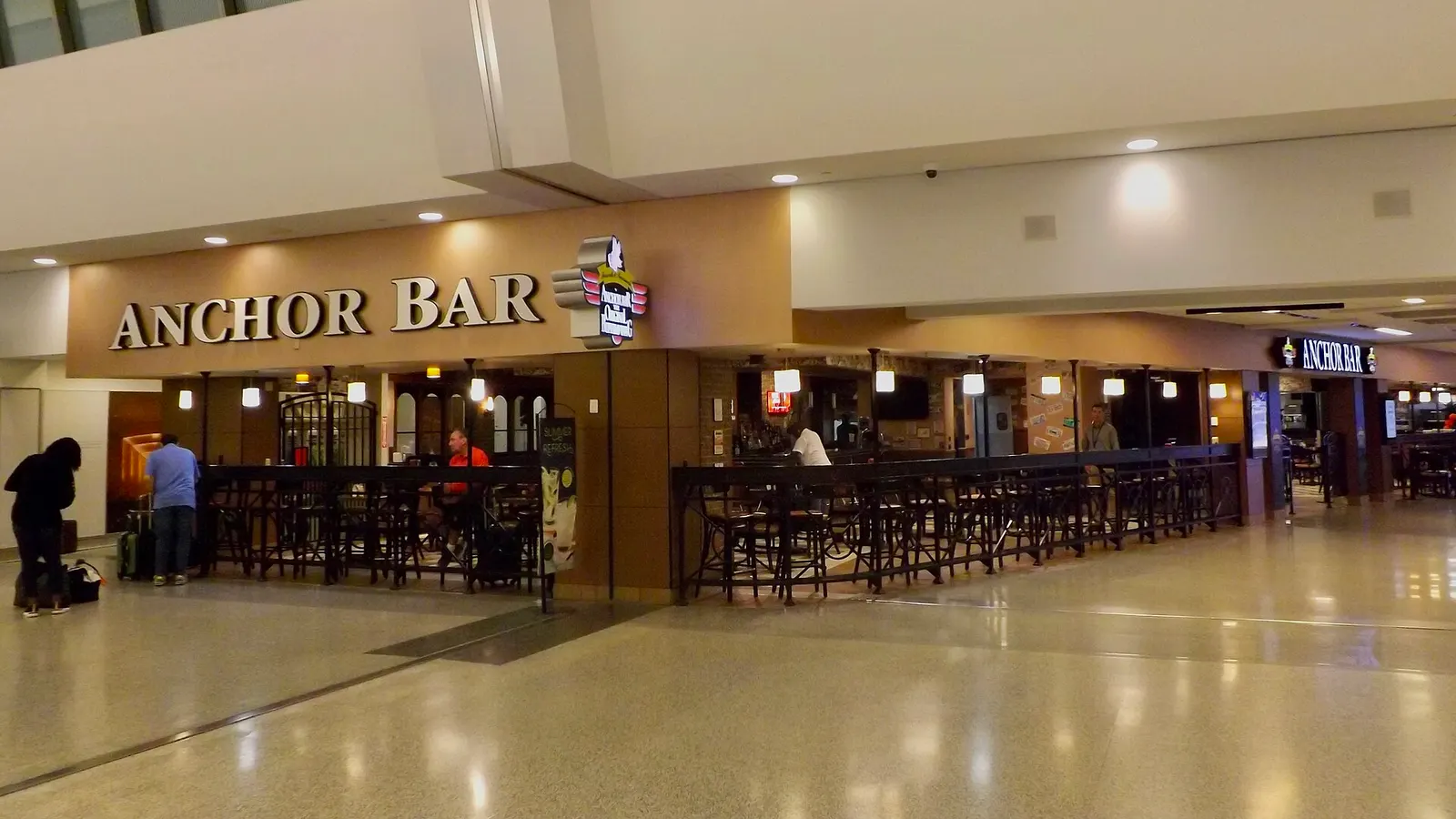 Airside Anchor Bar on the BUF concourse with Home of the Original Buffalo Wing branding, dark-wood bar, and travelers walking the terrazzo.