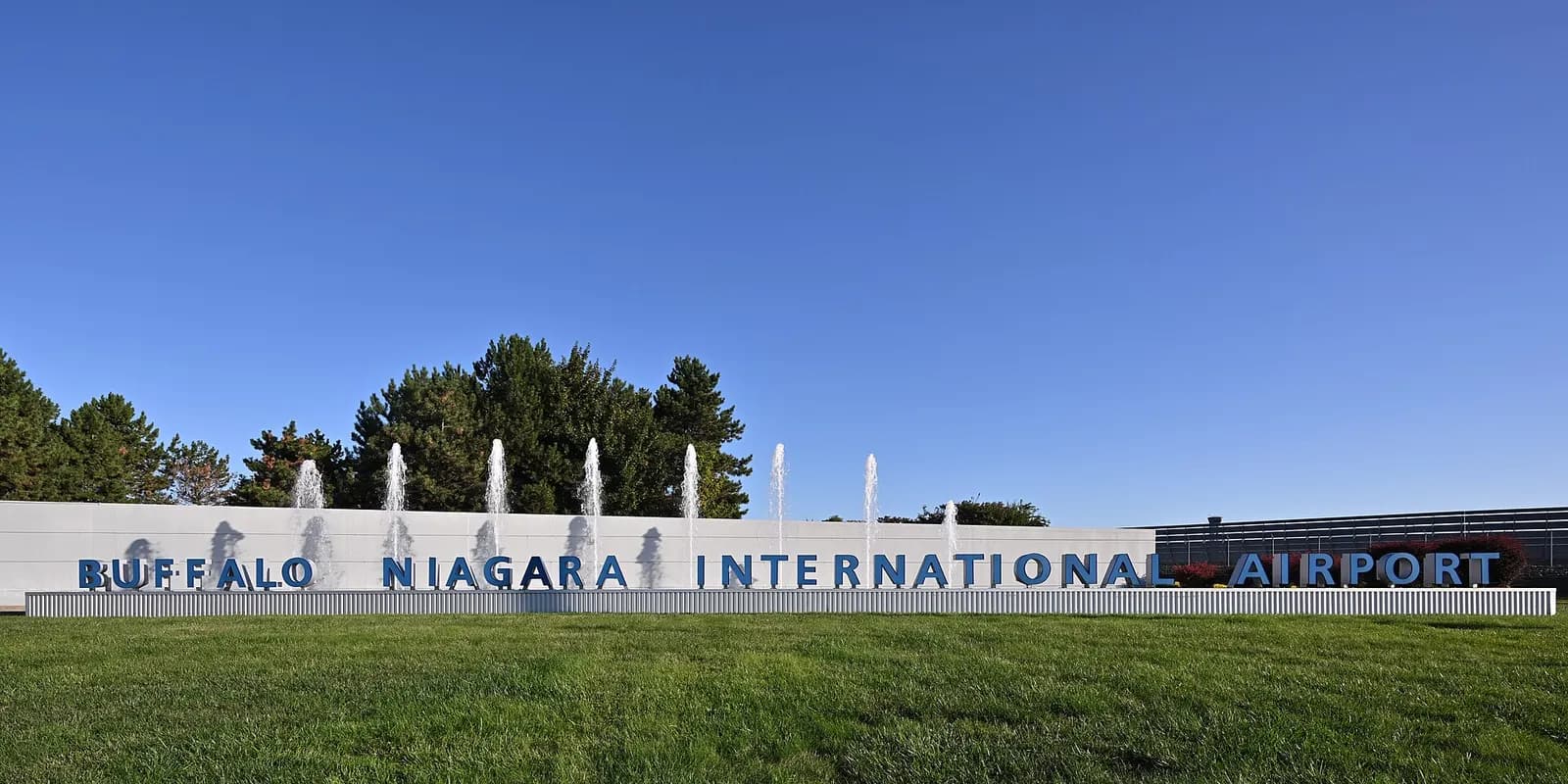 Blue three-dimensional letters spelling BUFFALO NIAGARA INTERNATIONAL AIRPORT in front of fountain jets and a white wall under a clear sky.