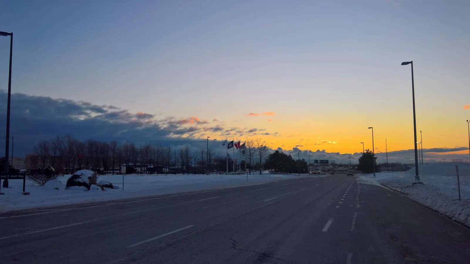 Snowy Luiz F. Kahl Way at BUF with the street sign in the foreground and U.S. and Canadian flags along the airport entrance at sunset.