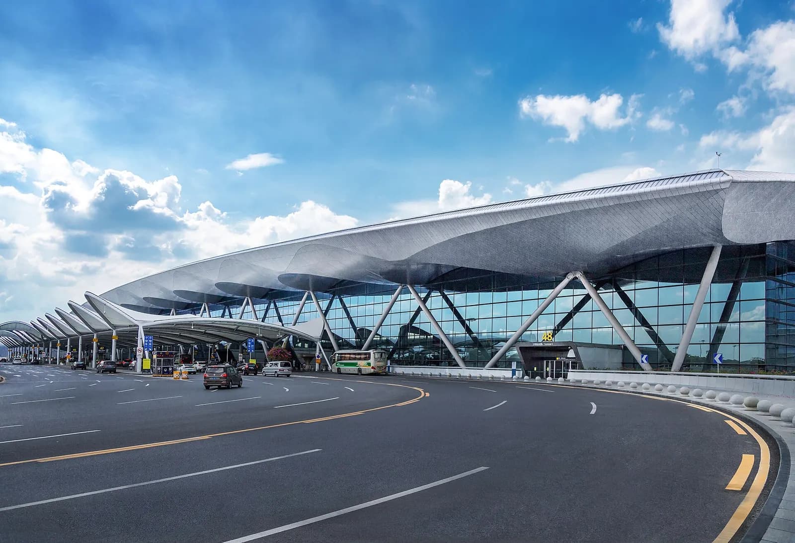 The glass-fronted main entrance of Terminal 2 with vehicle drop-off lanes, landscaping, and the airport name above the doors.