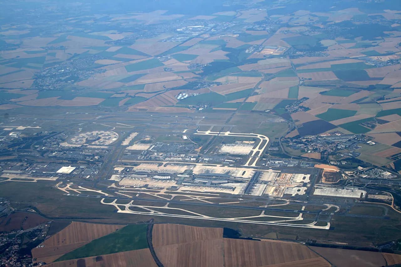 An aerial view of Charles de Gaulle Airport showing sprawling terminals, parallel runways, and taxiways across the Roissy countryside.