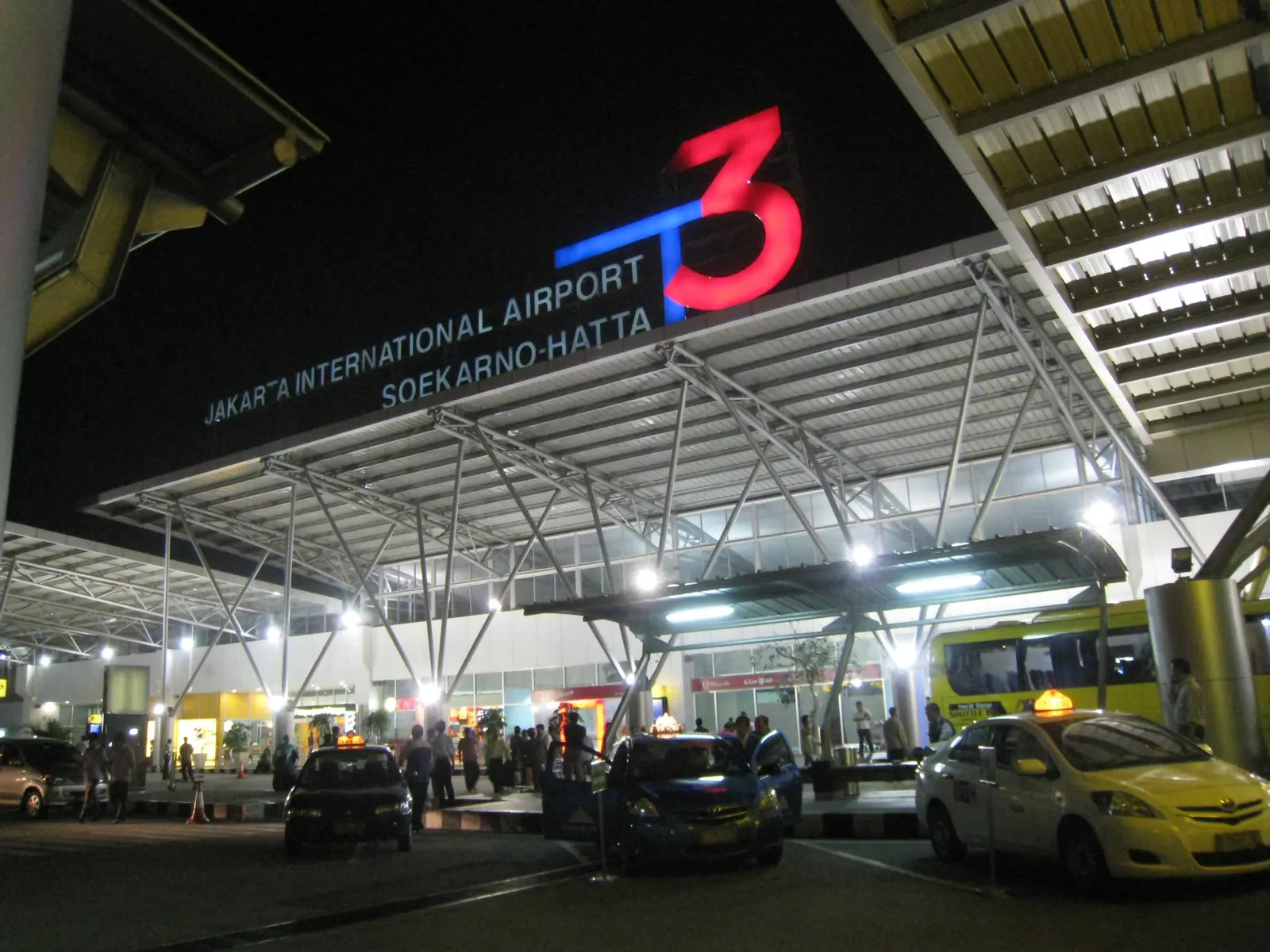 Curbside taxi and pickup lane in front of Terminal 3, with the terminal entrance canopy, roadway markings, and vehicles along the curb.