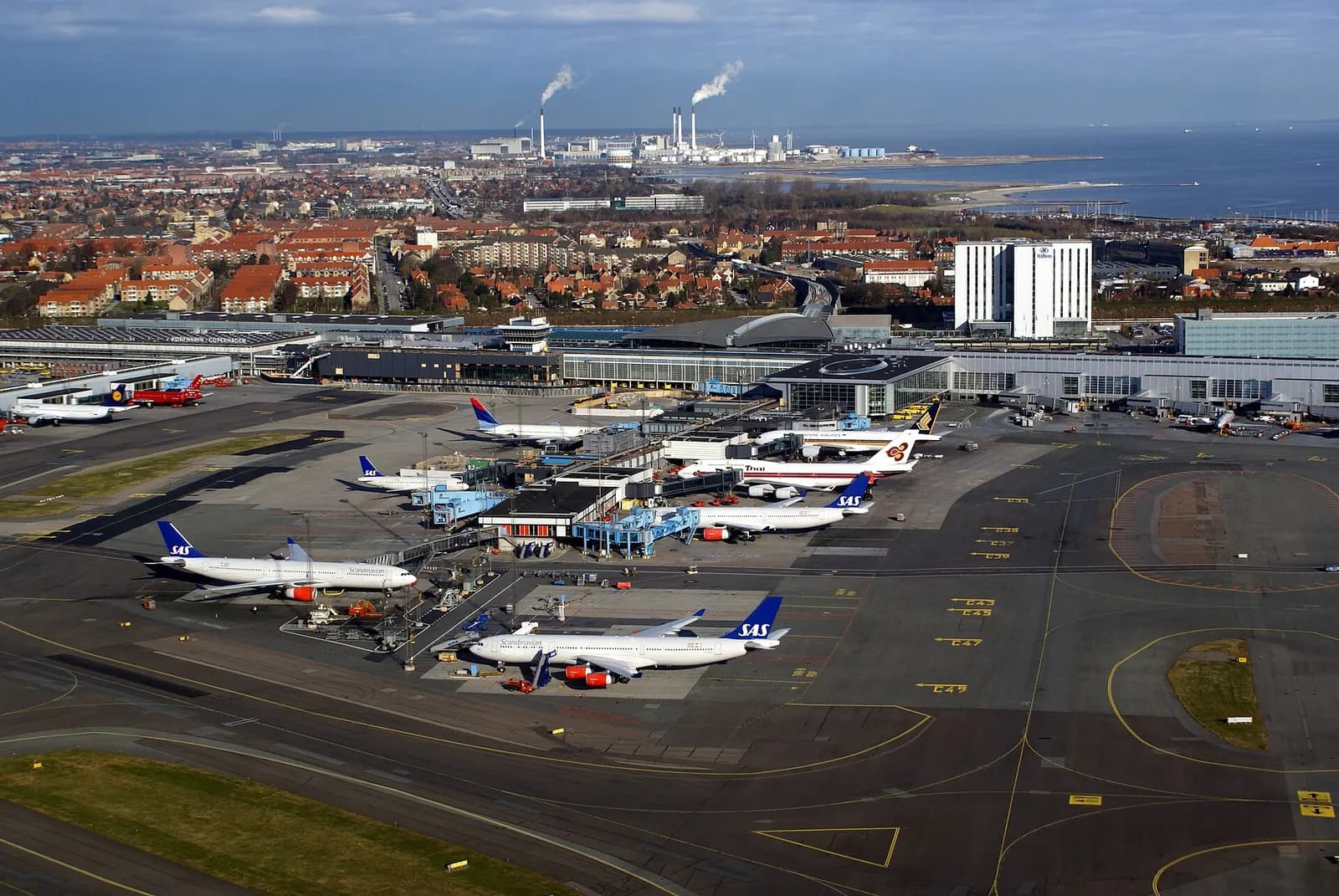 An aerial view of Copenhagen Airport showing Terminals 2 and 3, pier fingers, and parked aircraft.