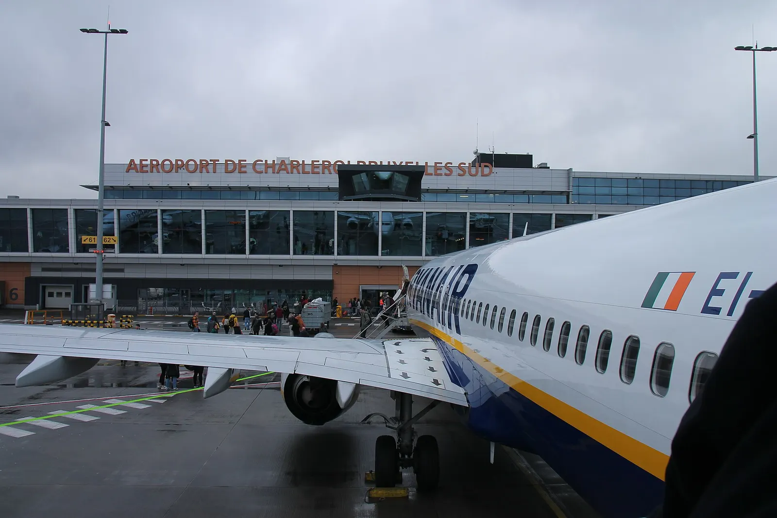 Ryanair Boeing 737 parked on a wet Charleroi stand with the glass terminal and orange airport lettering behind it.