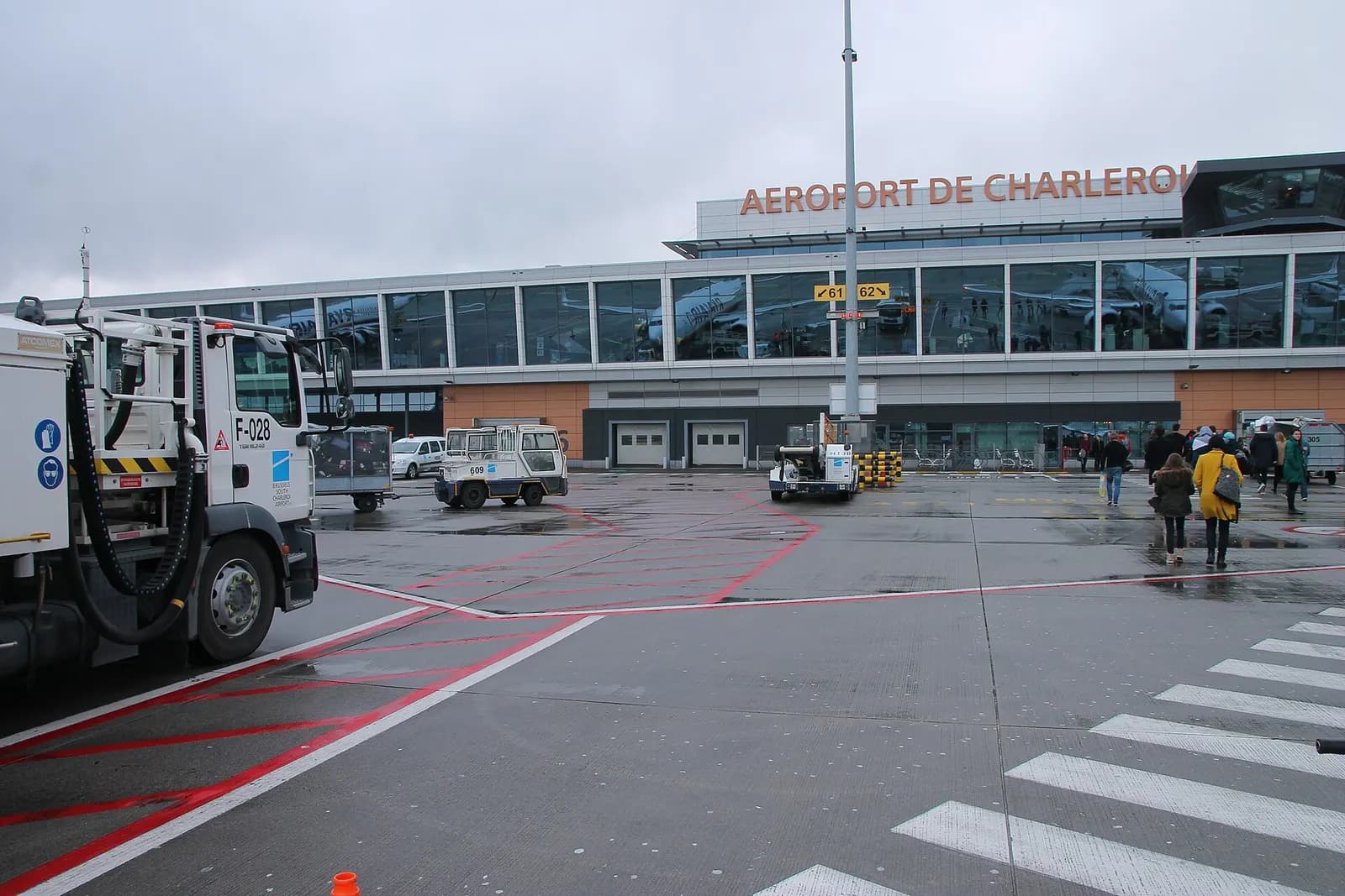 Passengers crossing the zebra path toward Charleroi’s glass terminal while a branded airport service truck sits on the wet apron.
